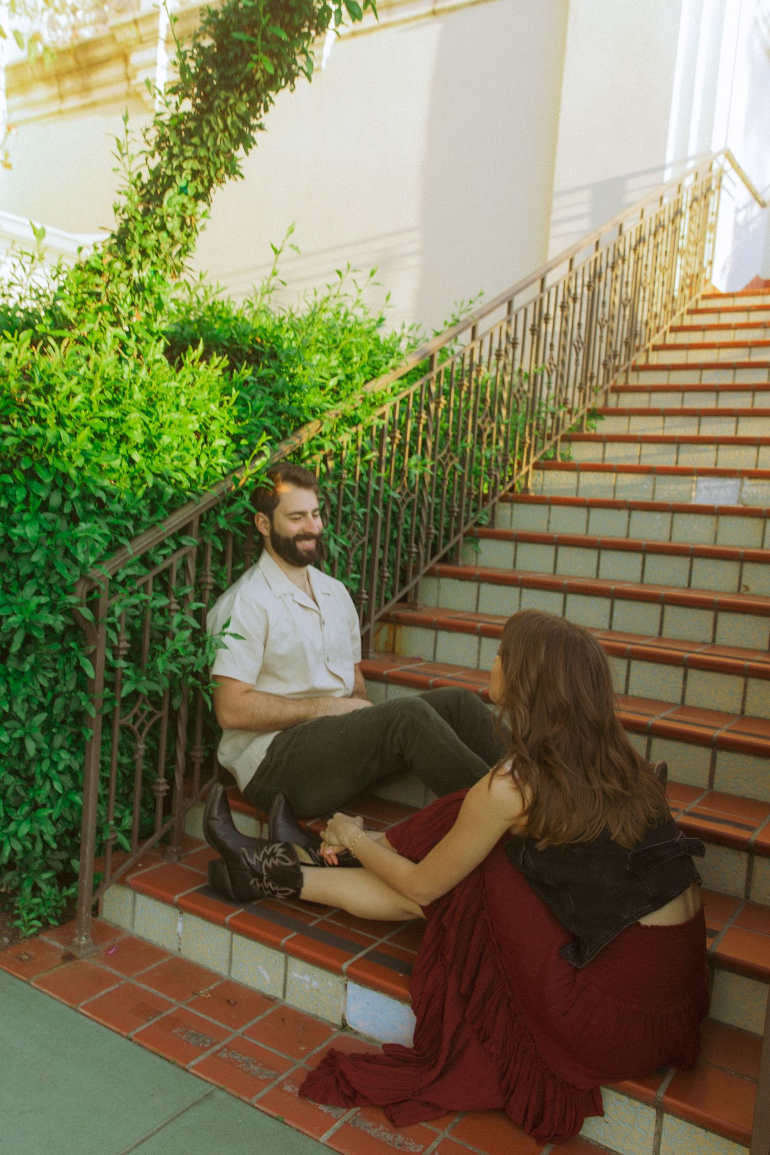 A couple shares a relaxed moment on outdoor brick stairs with lush greenery and wrought iron railings.