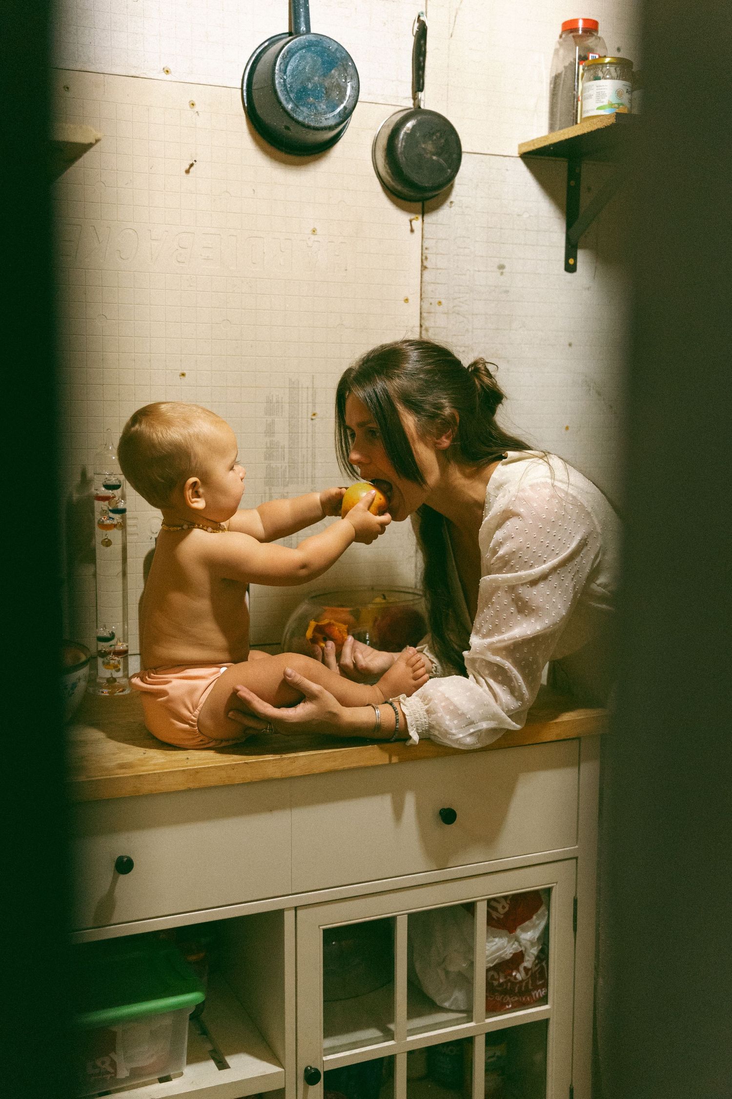 Intimate parenting moment captured through window frames in a dimly lit nursery setting.