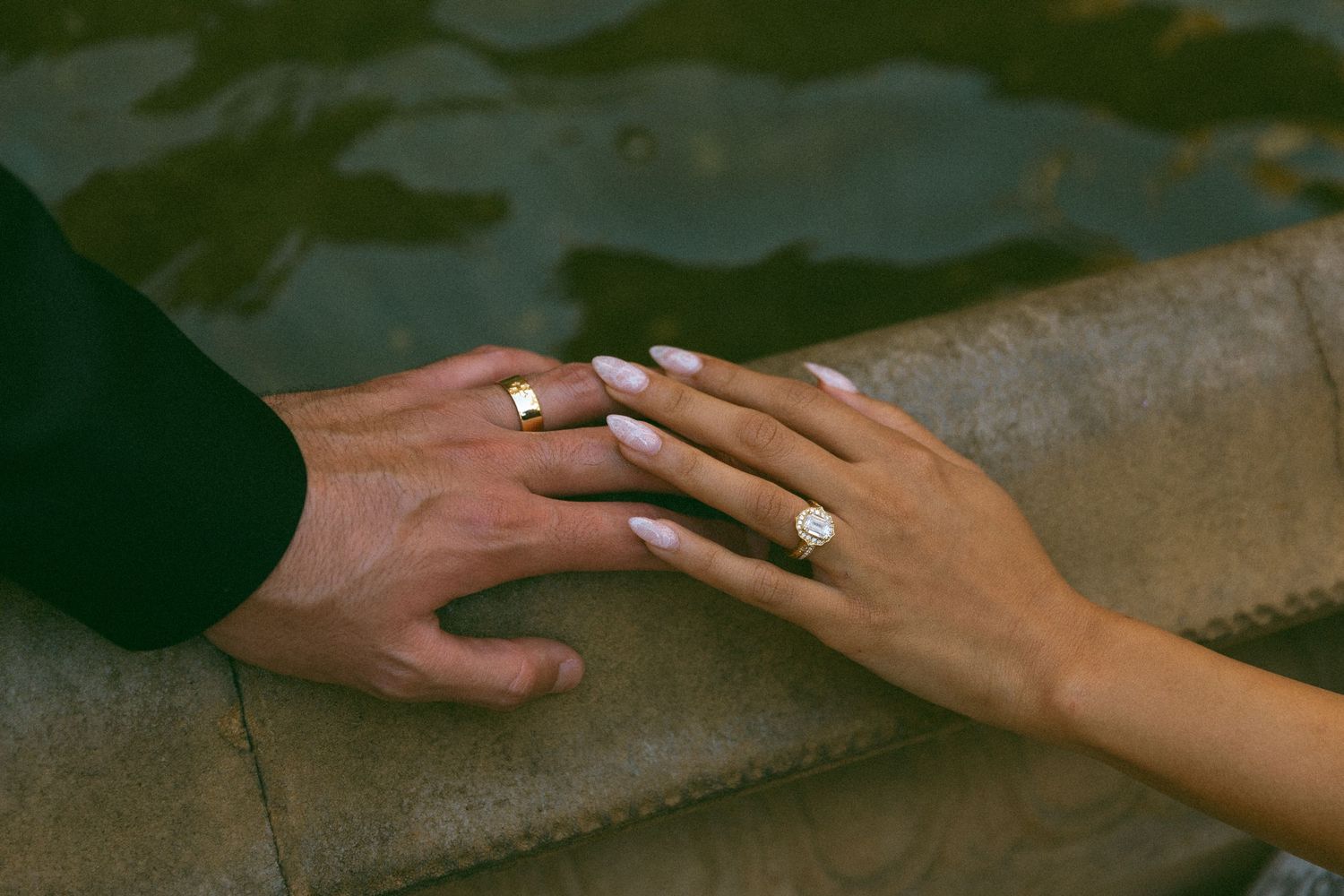 Couple's hands resting together on a stone ledge by water, her manicured hand showing a diamond engagement ring beside his gold band.