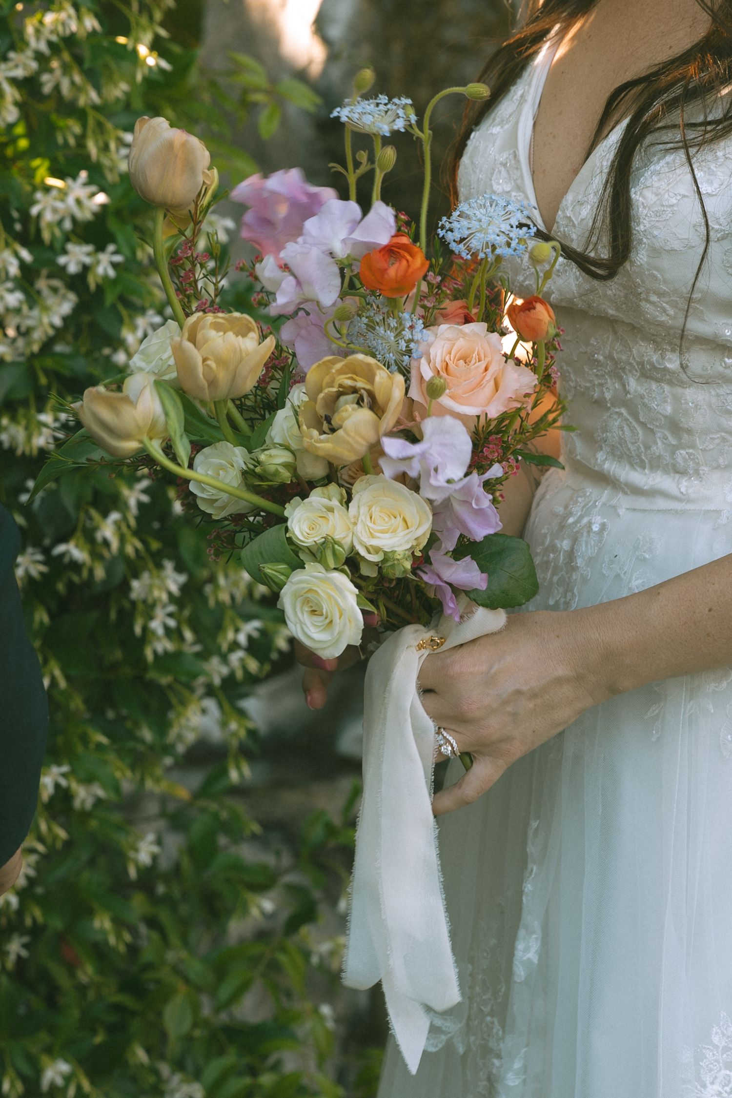Bride in lace wedding dress holds a lush bridal bouquet of peach, yellow, and pink roses with white ribbon.