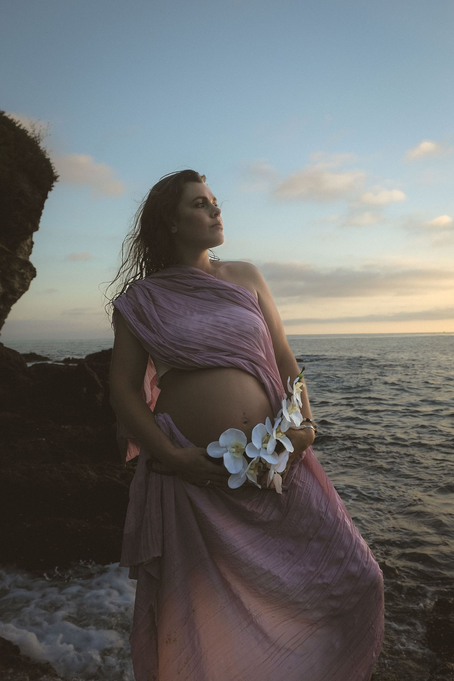 Dramatic oceanside maternity portrait with flowing purple dress at golden hour.