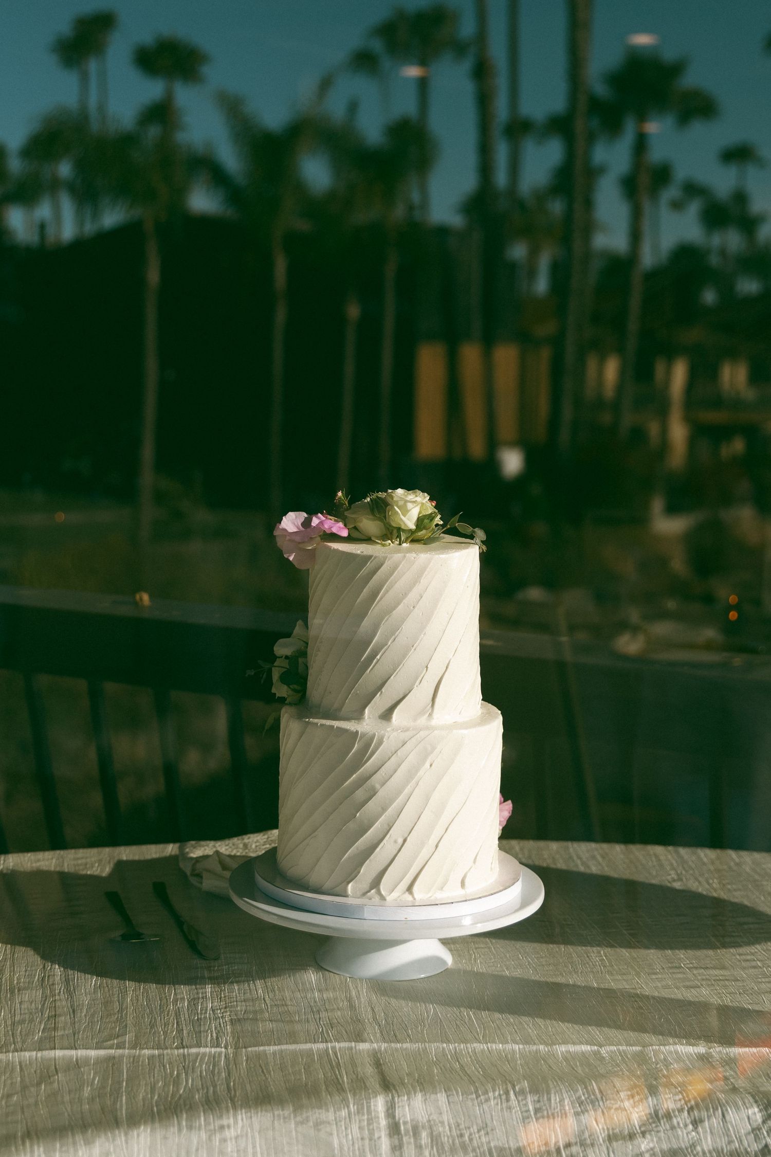 Elegant two-tier white wedding cake with diagonal texture and pink flowers on a table outdoors.
