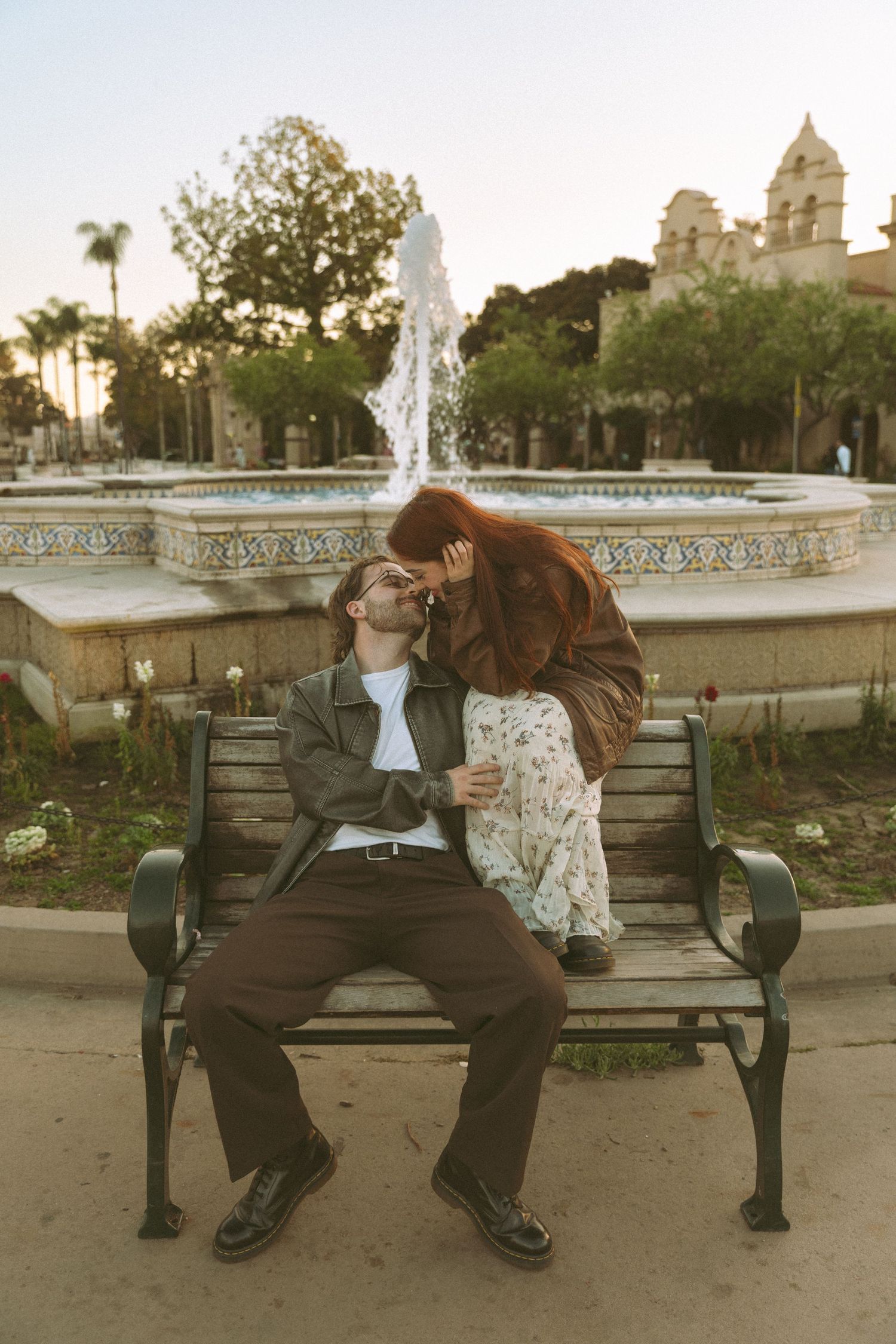 Couple sharing tender moments on park bench with fountain and architecture in background.