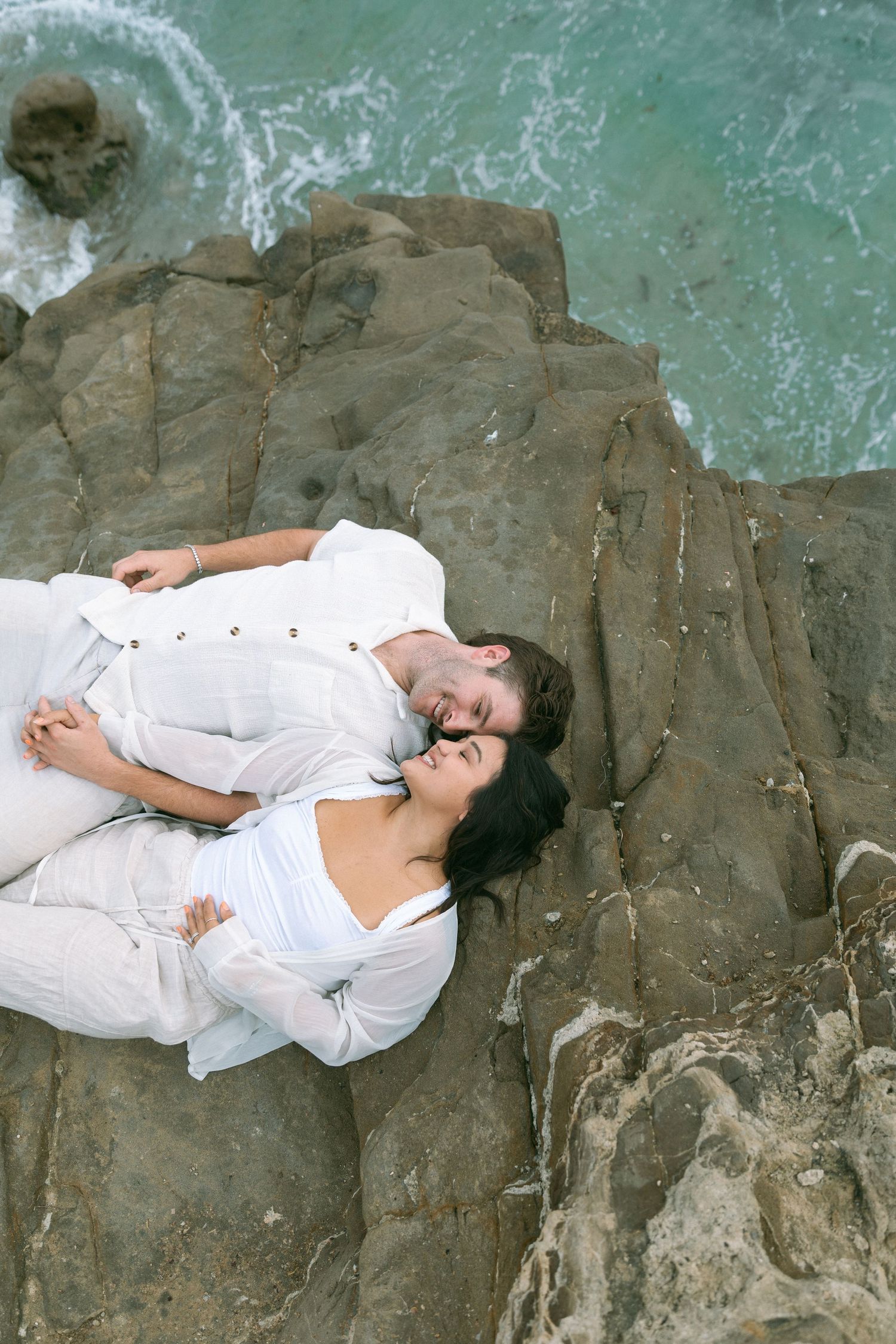 Two people in white clothing lay on coastal rocks near turquoise waters.