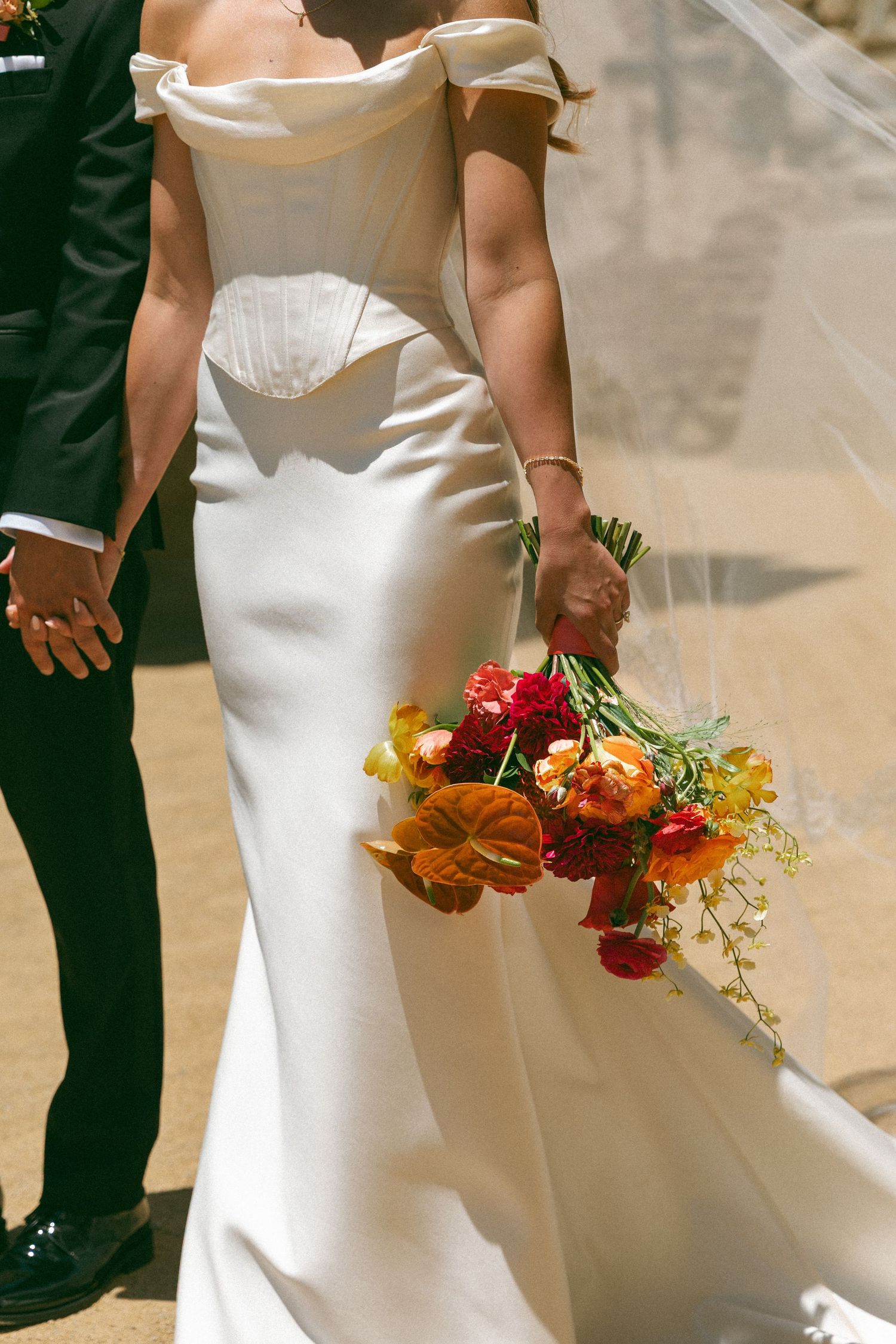 Bride in off-shoulder white wedding dress holding vibrant red and orange bouquet while walking with groom.