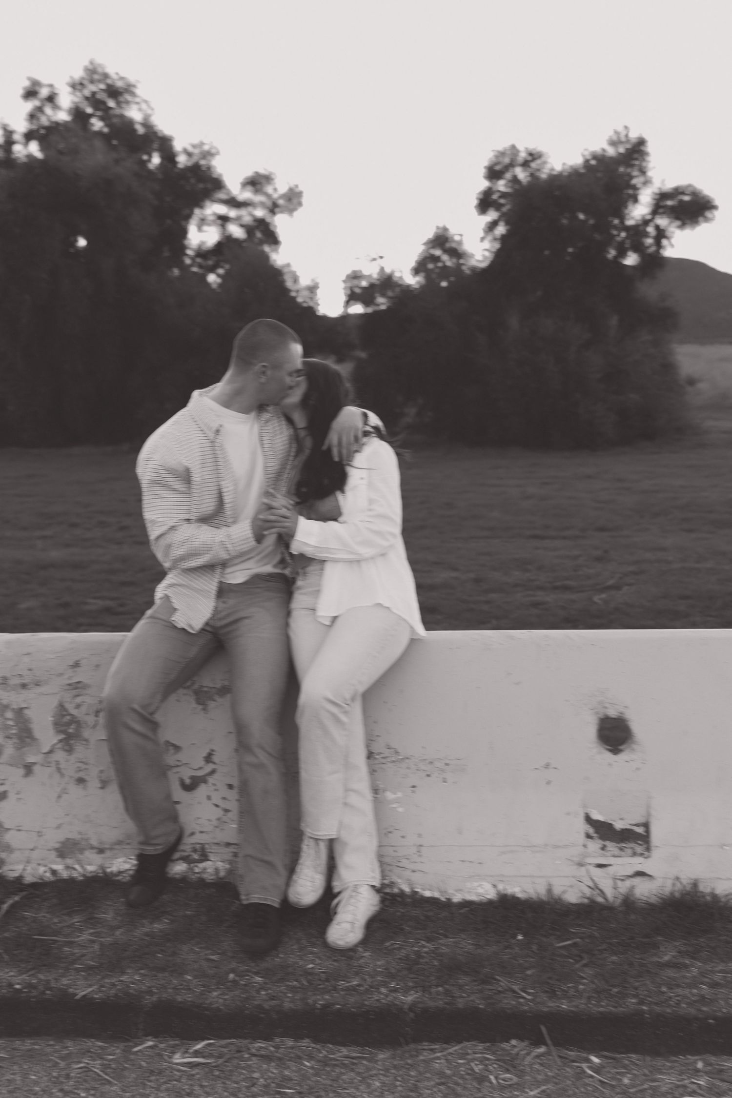 A couple in white outfits sharing a tender moment against a concrete wall at dusk.