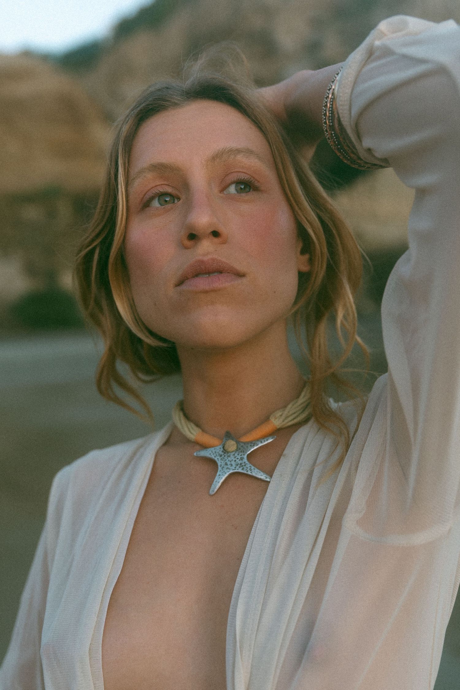 Portrait shot in warm light showing person wearing white flowing top and starfish pendant necklace against blurred outdoor background.