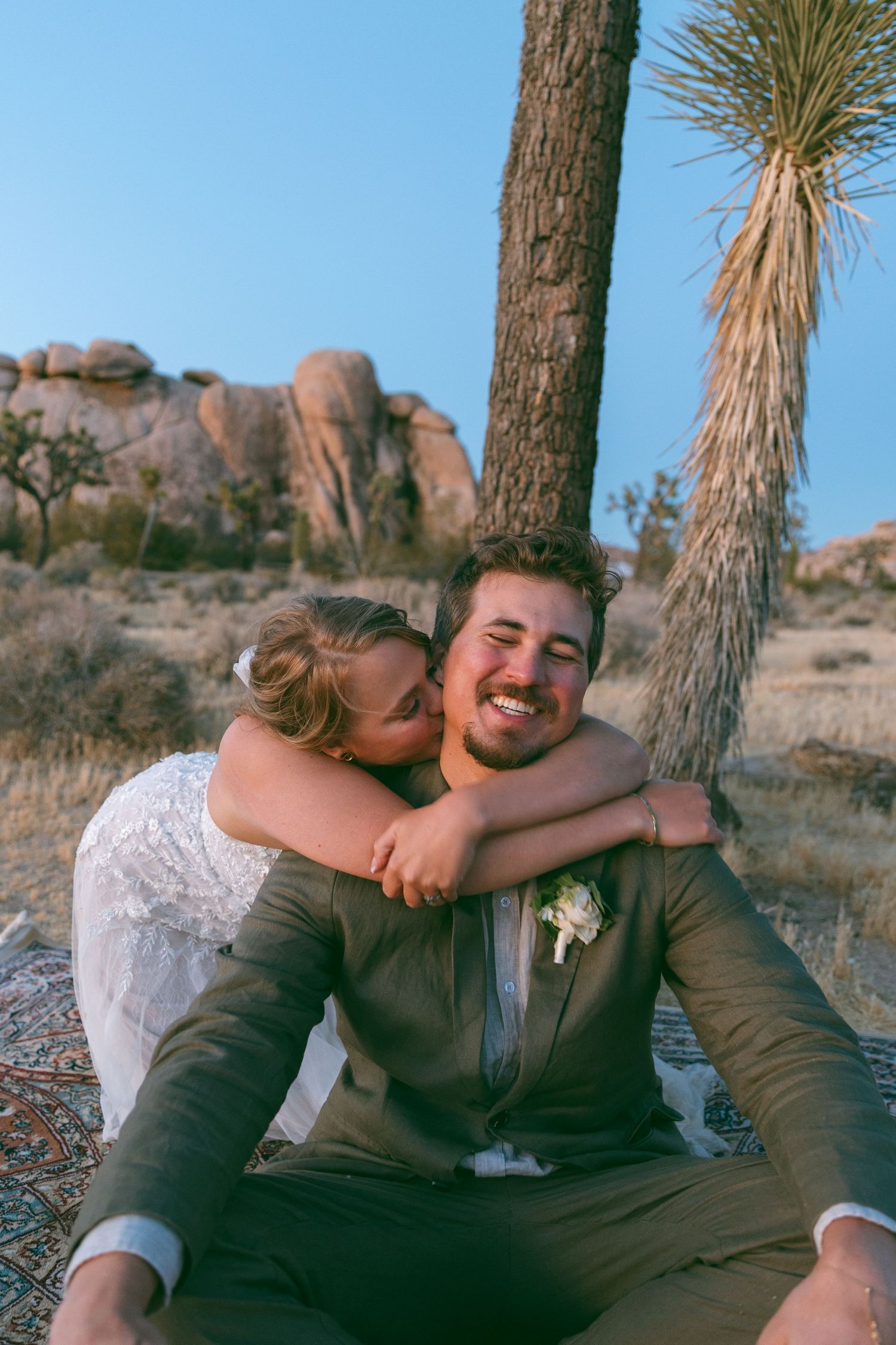A bride embraces a smiling groom from behind as they sit together in Joshua Tree desert landscape.