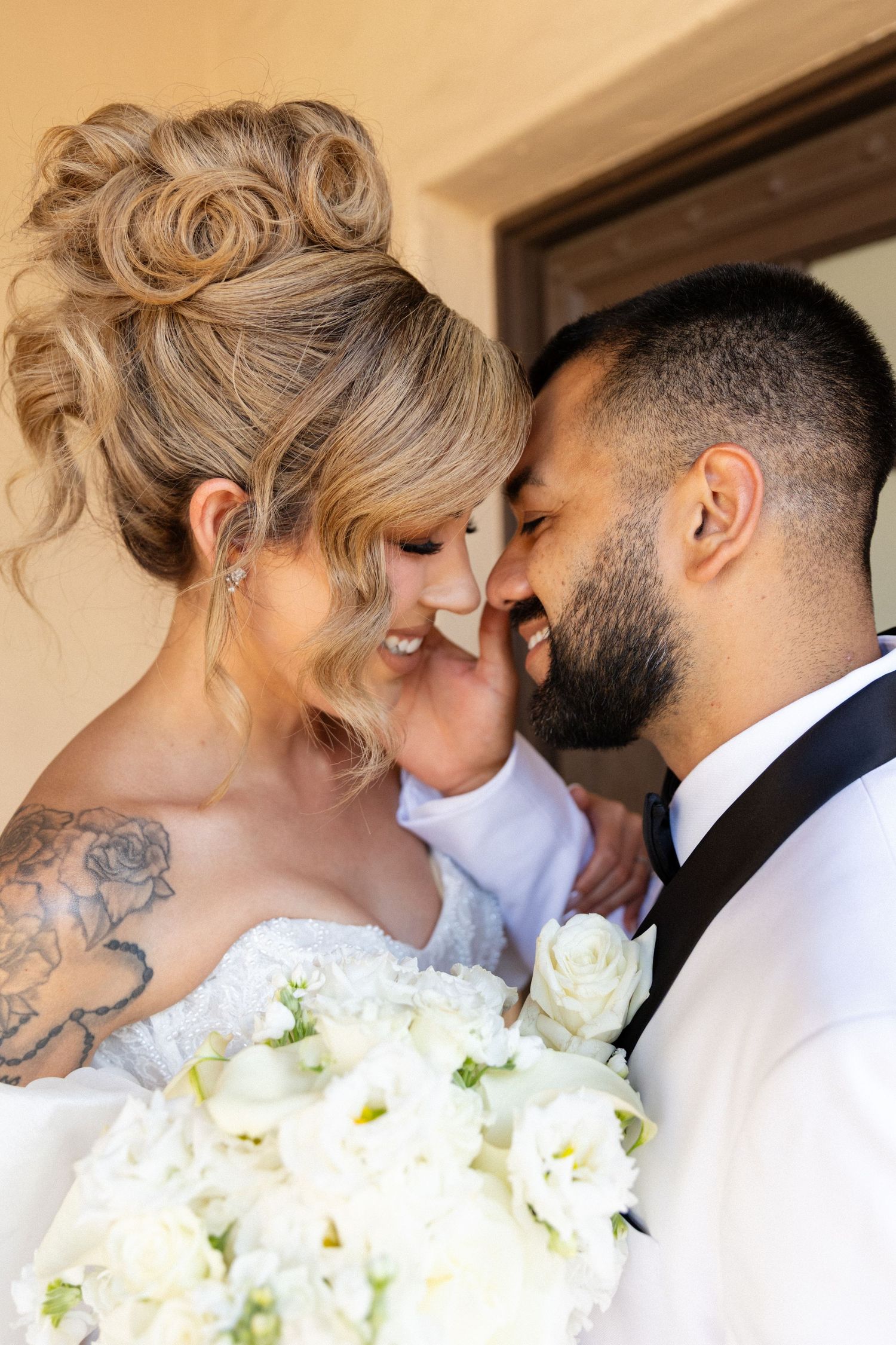 Bride and groom share intimate moment, bride holding white rose bouquet with elegant updo and tattoo.