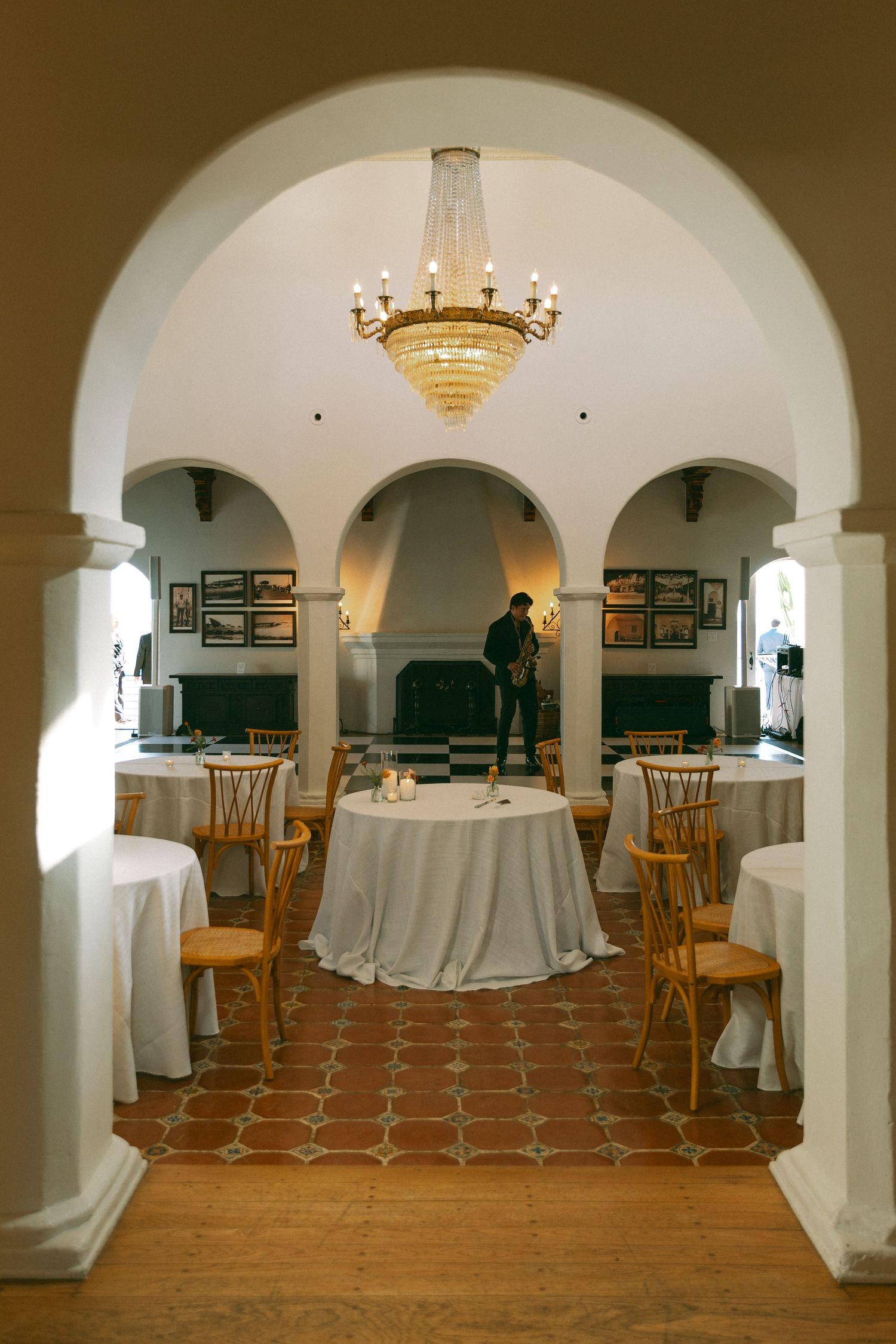 Elegant wedding venue interior with arched doorways, crystal chandelier, round tables, and terracotta tile flooring.