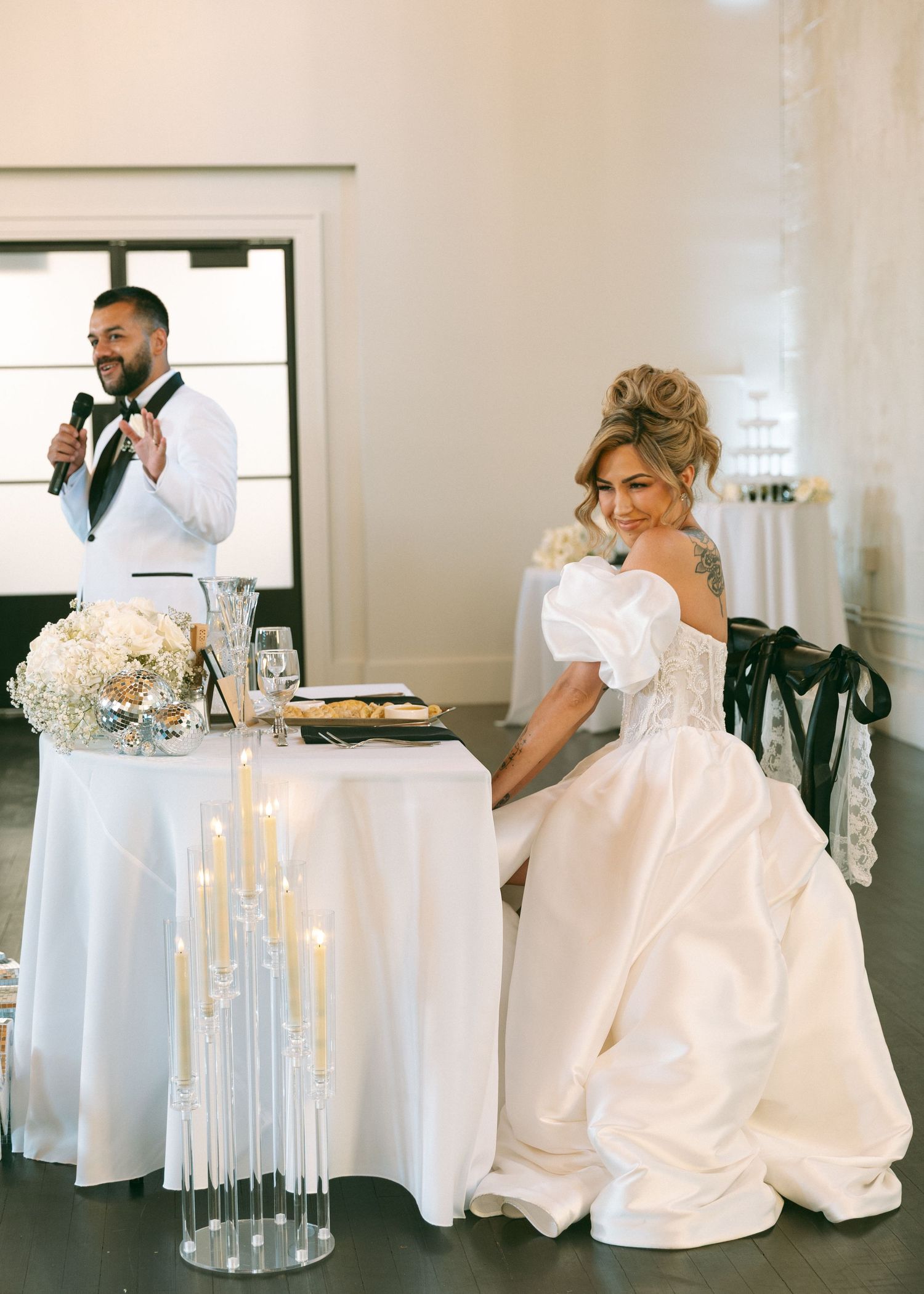 Groom speaks at wedding reception while bride sits at sweetheart table with candles.