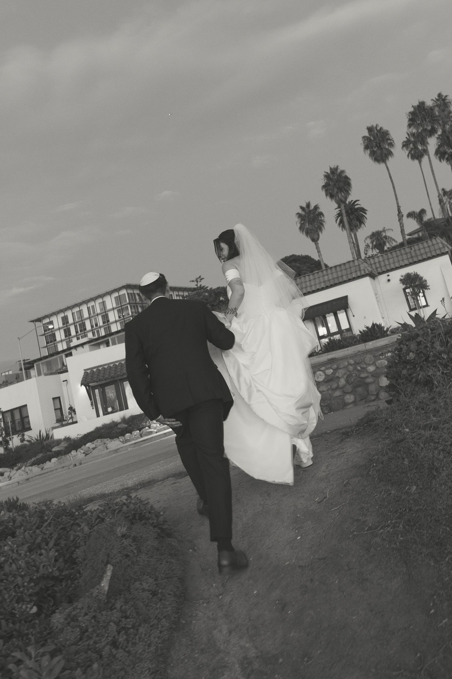 A wedding couple dances together on a grassy hill with palm trees and buildings in the background.