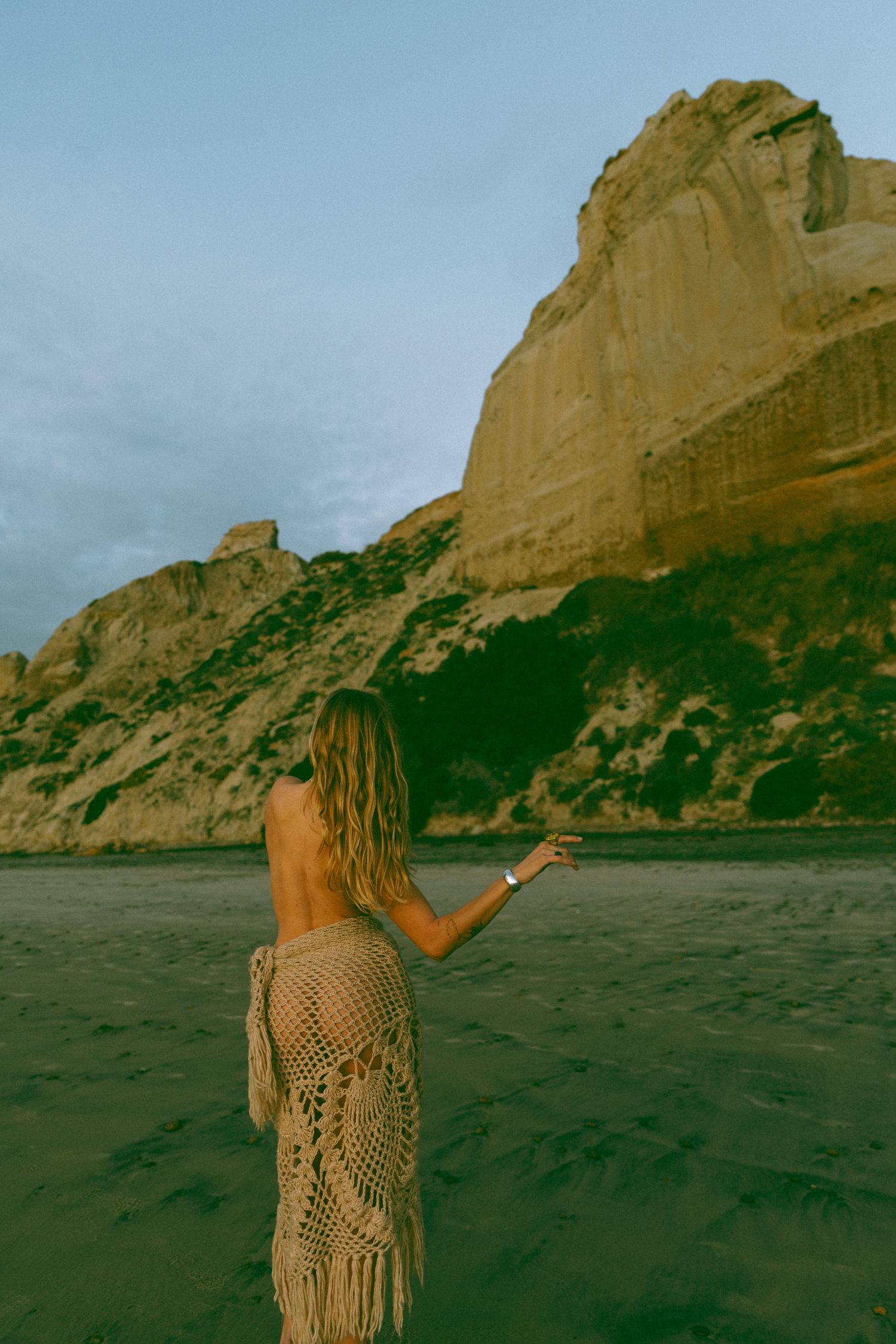 A person in a crochet dress stands on a beach gesturing towards towering coastal cliffs during golden hour.