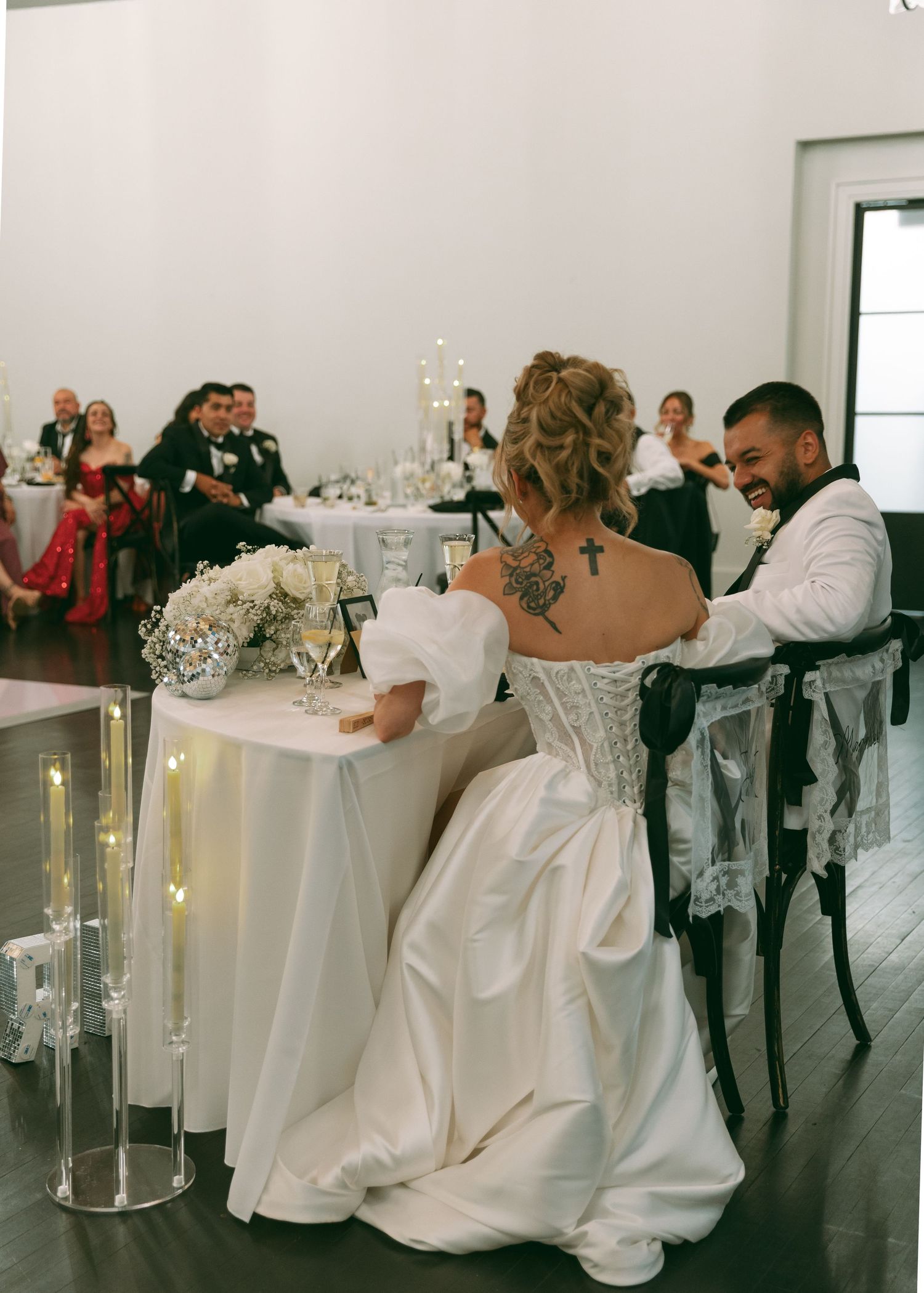 Bride and groom sit at sweetheart table during wedding reception, bride showing back tattoo.