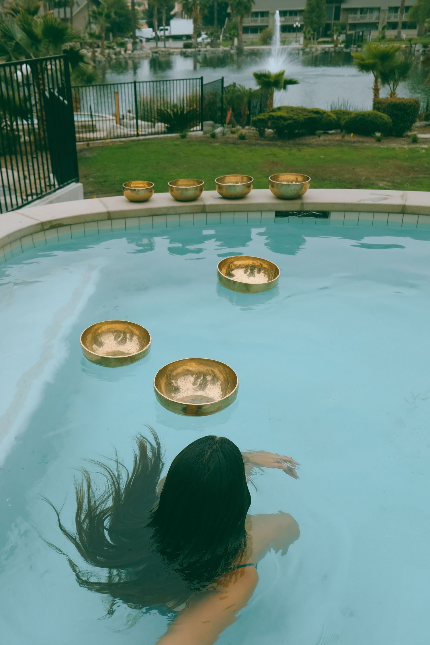 Golden singing bowls float in a blue pool with palm trees and fountain in the background.