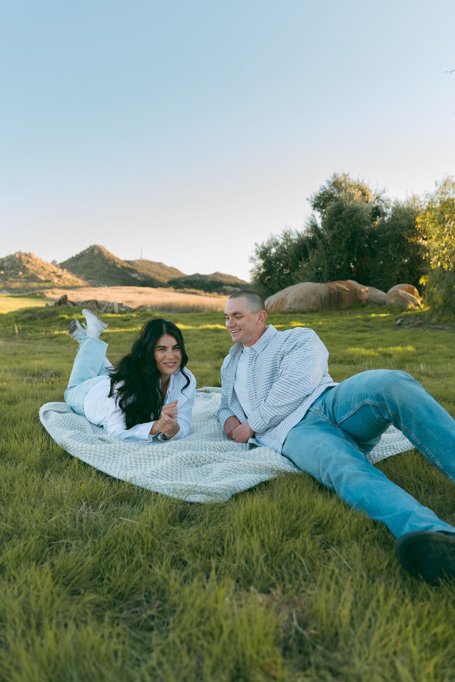 A couple relaxes on a picnic blanket in a grassy field with rolling hills in the background.