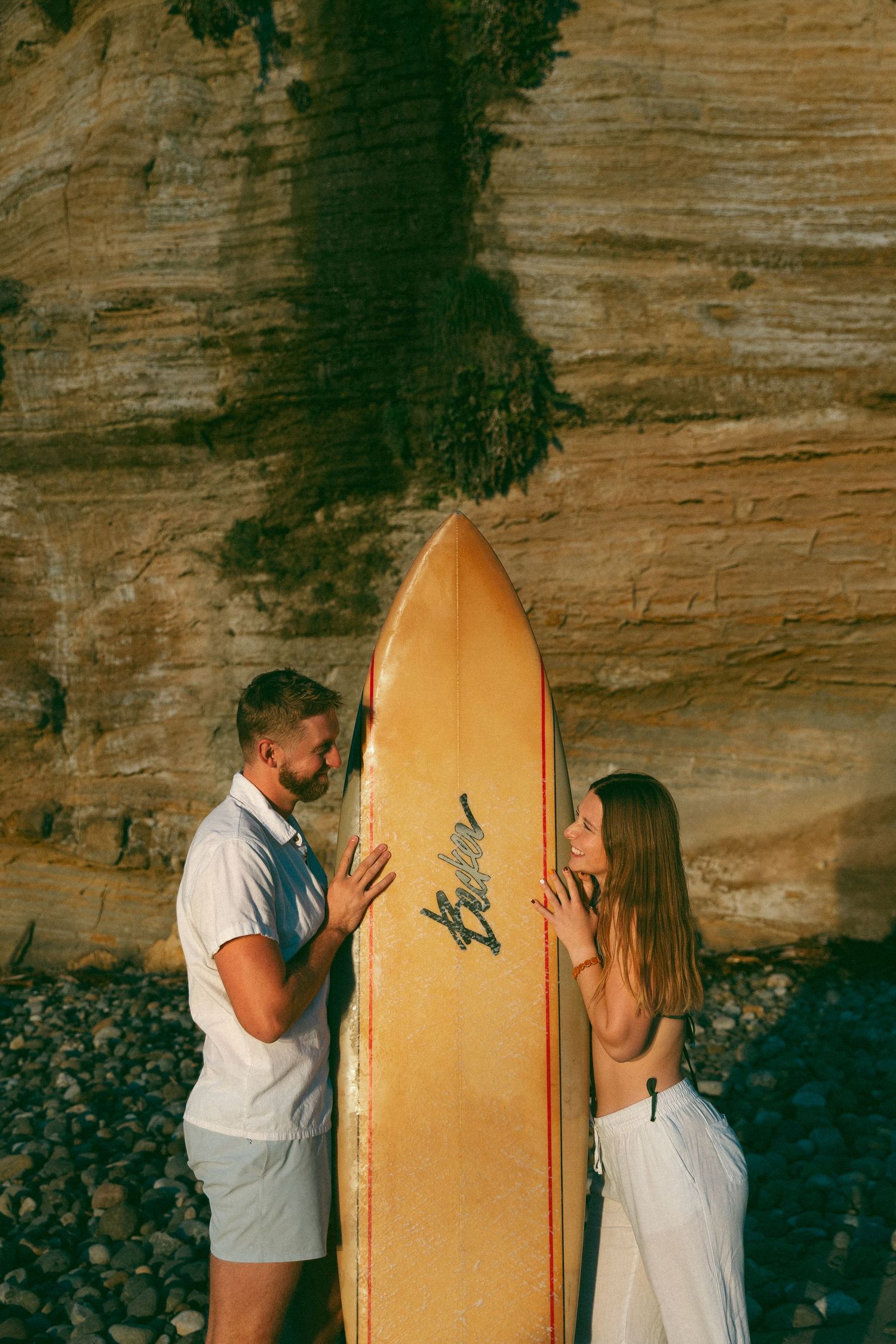 A couple playfully poses with a yellow surfboard against a rocky cliff backdrop.