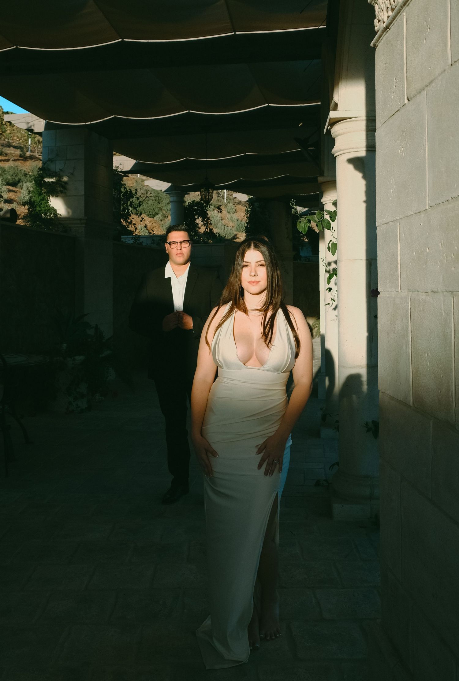 A couple in formal attire poses in a shadowy outdoor corridor with dramatic lighting.