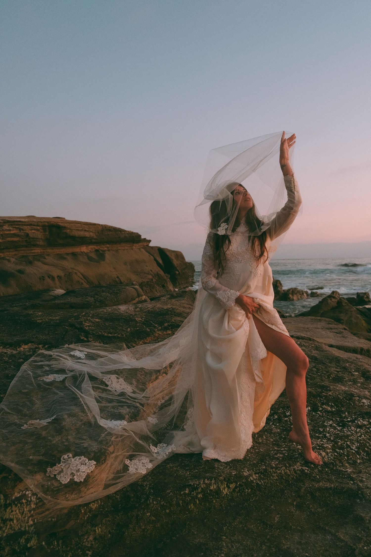 Bride in flowing white gown and veil standing on rocky coastal cliffs at sunset.