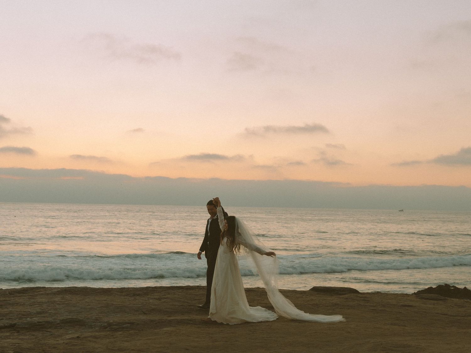 Bride and groom embrace on a sandy beach at sunset with ocean waves and pastel sky backdrop.