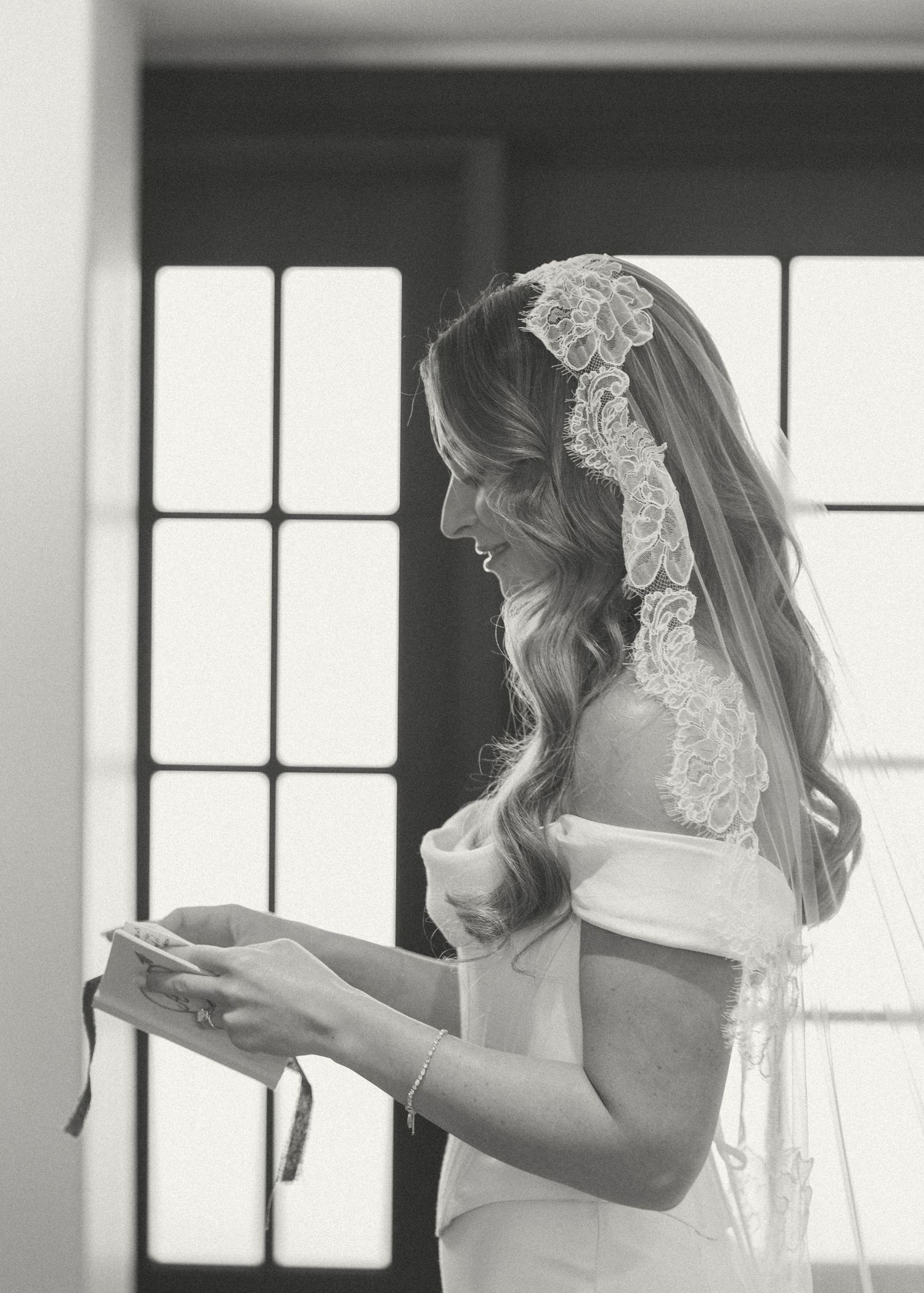Elegant bride in off-shoulder gown and lace mantilla veil reads vows by window in black and white photo.