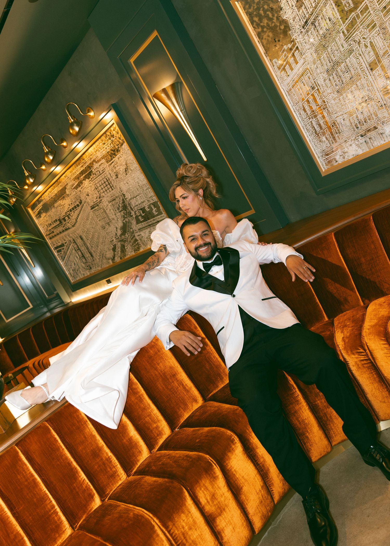 Bride and groom posing playfully on a velvet sofa in an elegant venue, groom in white tuxedo holding bride.
