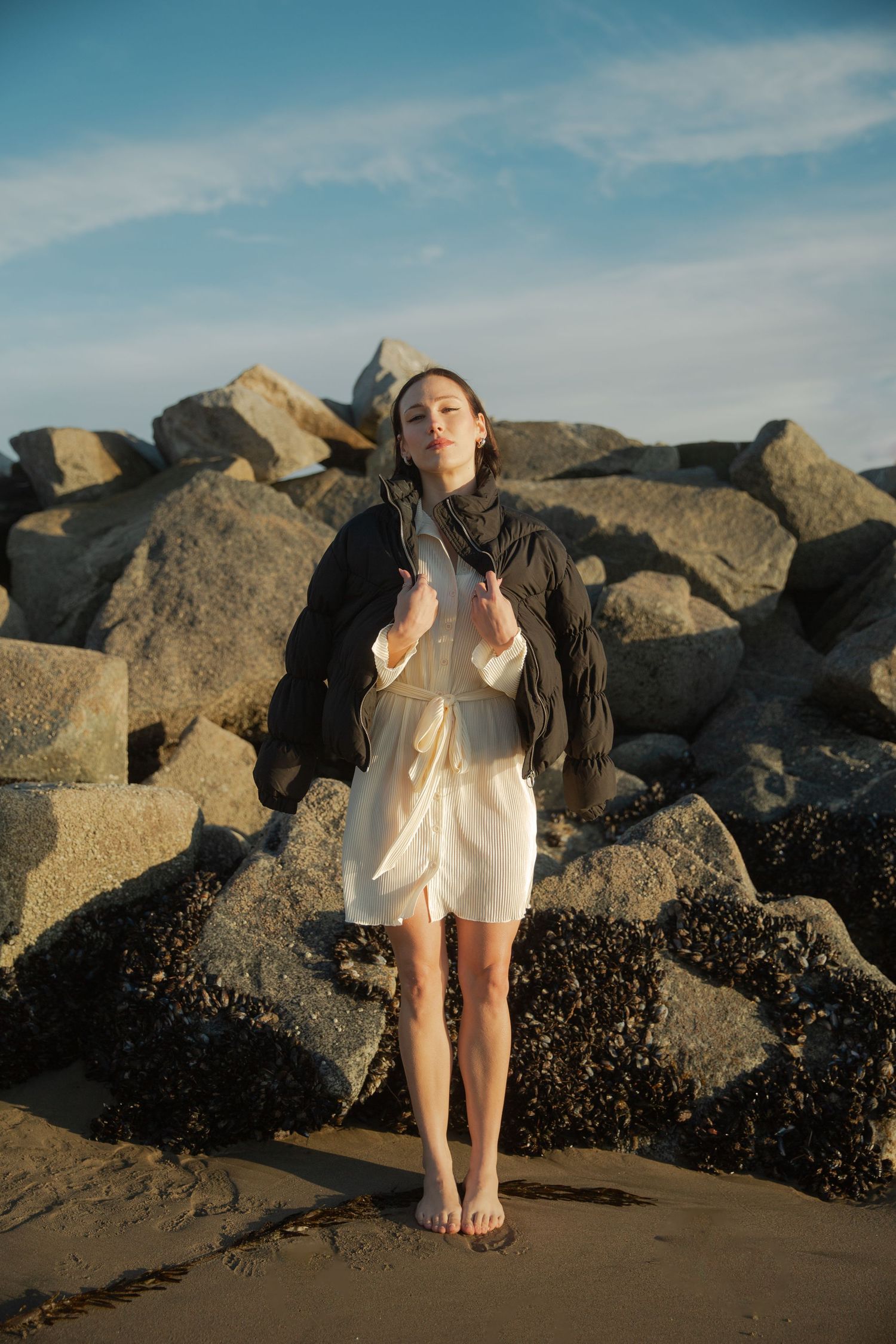 Figure in white dress posed among coastal rocks during golden hour.