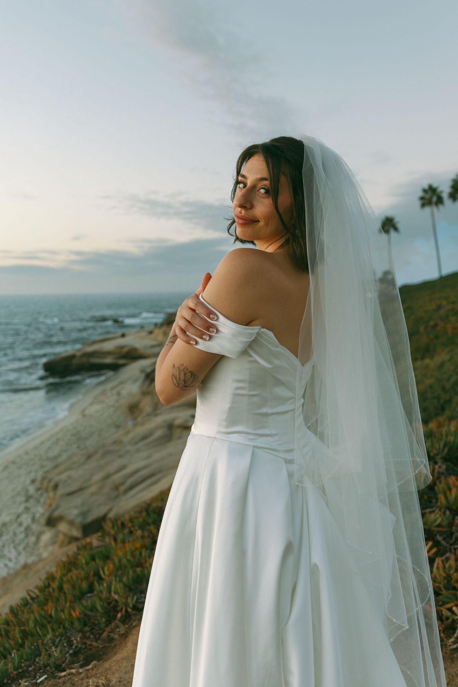 A bride in a white dress poses on a scenic coastal cliff overlooking the ocean at sunset.