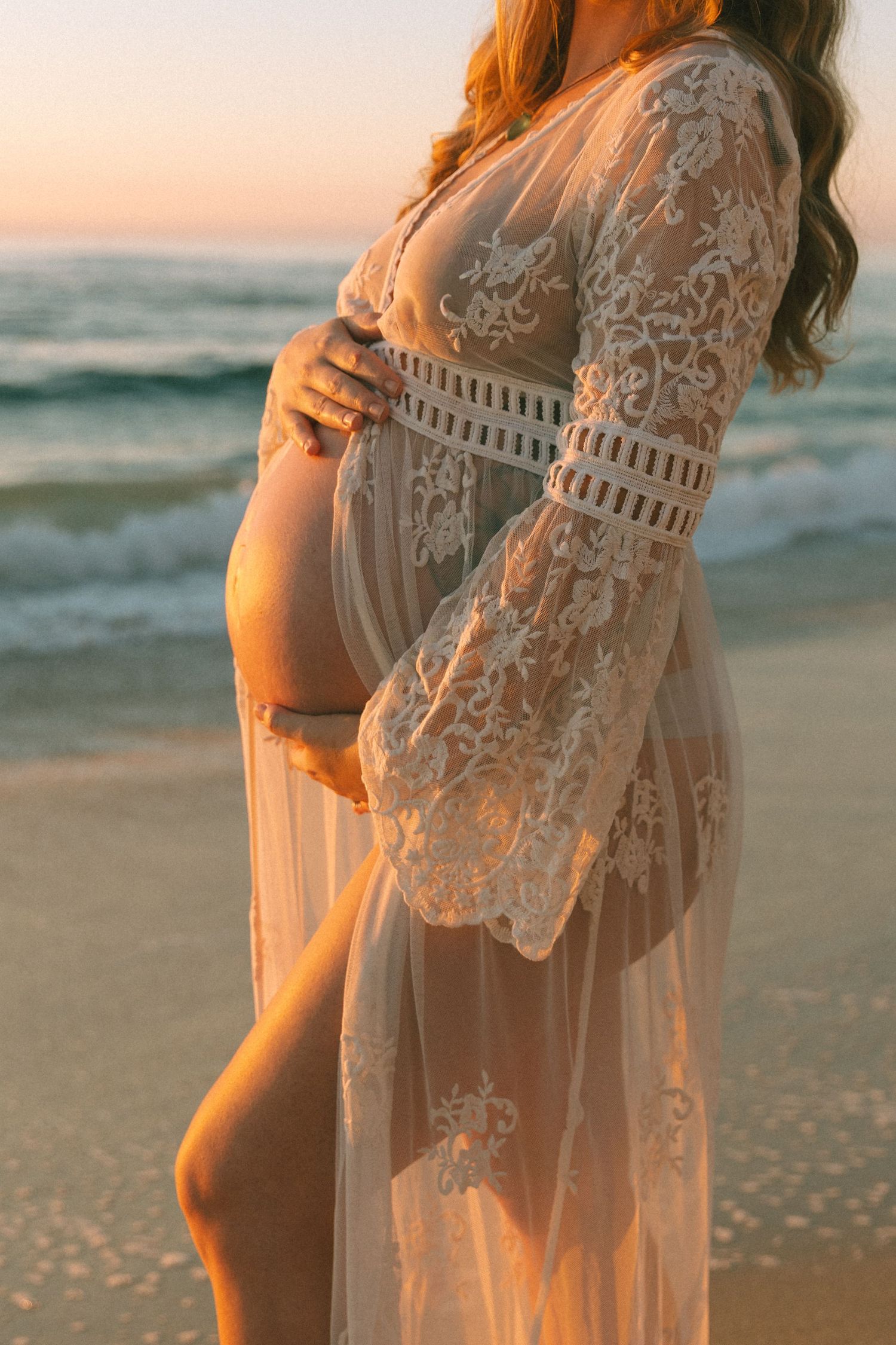 A flowing lace cover-up dress photographed against a sunset beach backdrop with waves in the background.