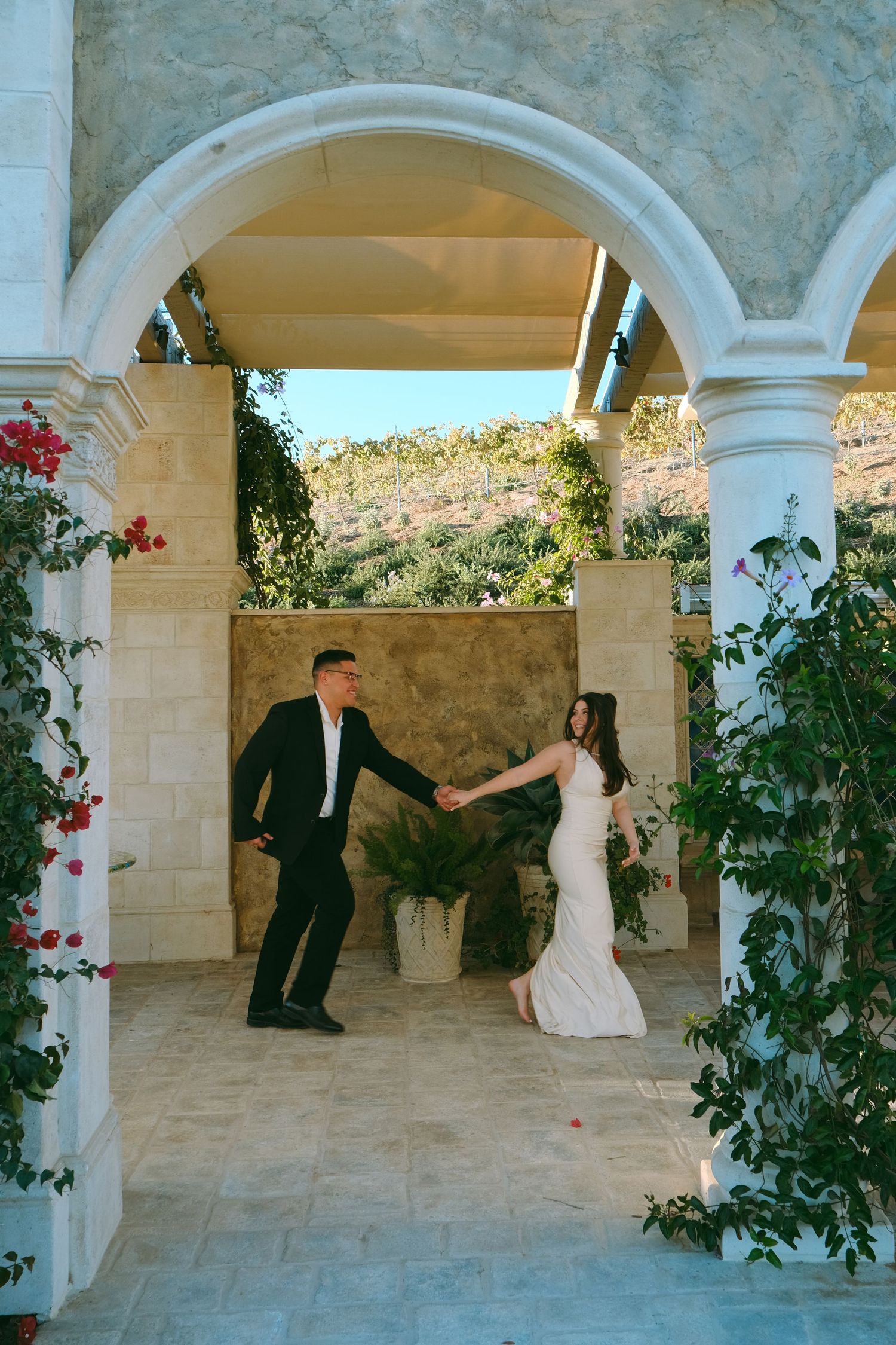 A couple dancing beneath an elegant stone archway surrounded by climbing flowers and greenery.