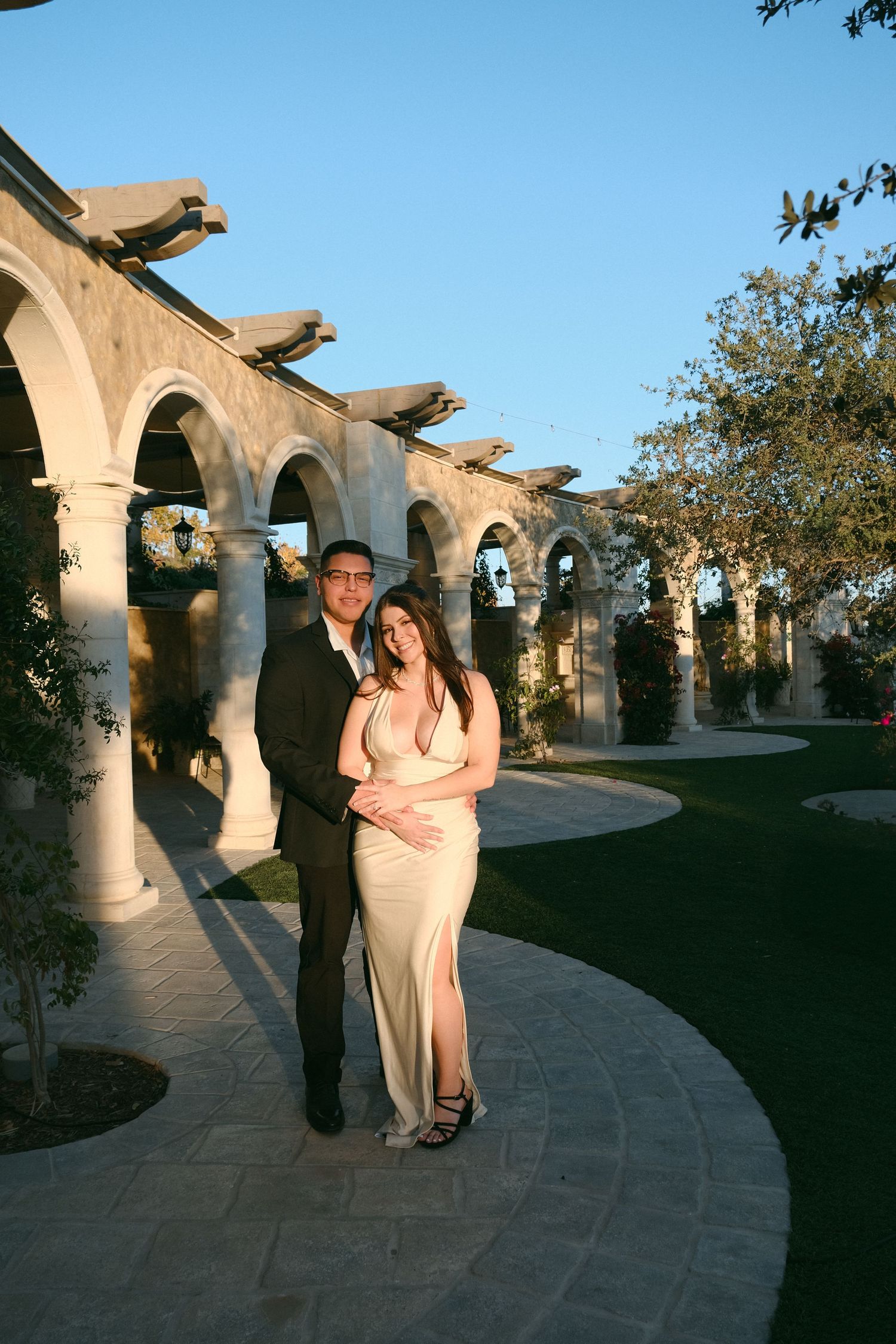 Couple embracing in formal attire along stone colonnade pathway at dusk.