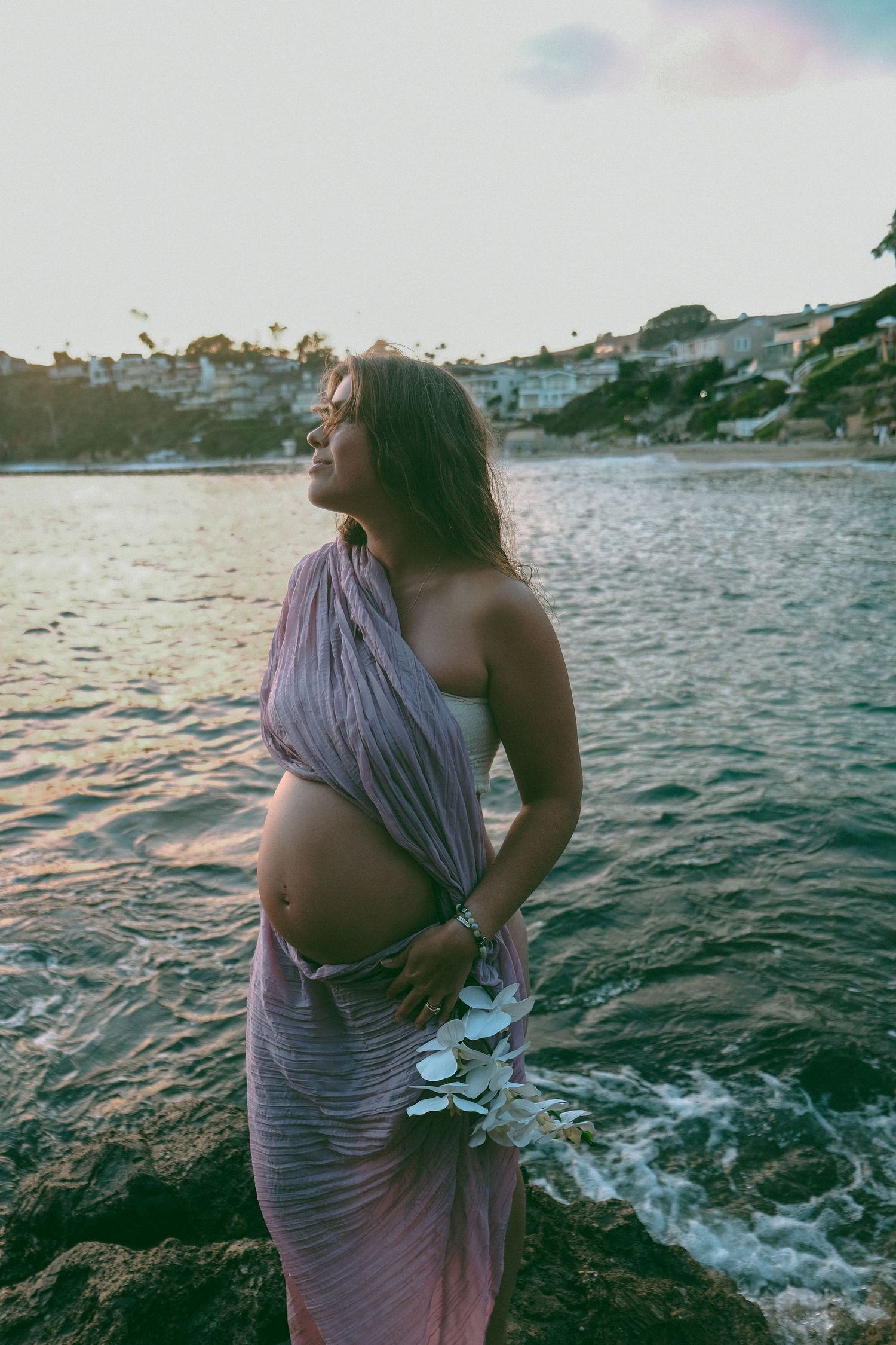 Maternity photoshoot at sunset by the ocean showing a draped purple dress against water and sky.
