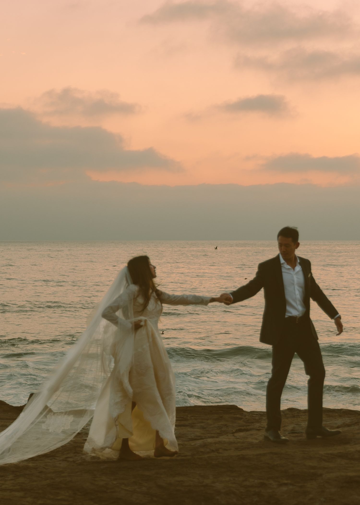 A bride and groom hold hands on a sandy beach at sunset, with a flowing veil and warm pink sky backdrop.