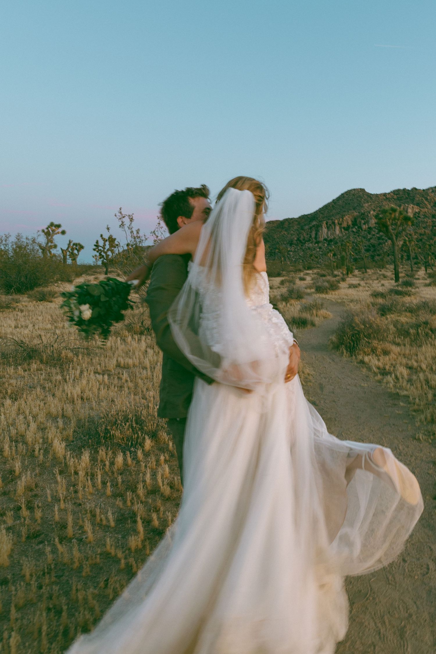 Bride in white gown and veil kissed by groom in desert landscape with Joshua trees and mountains at dusk.