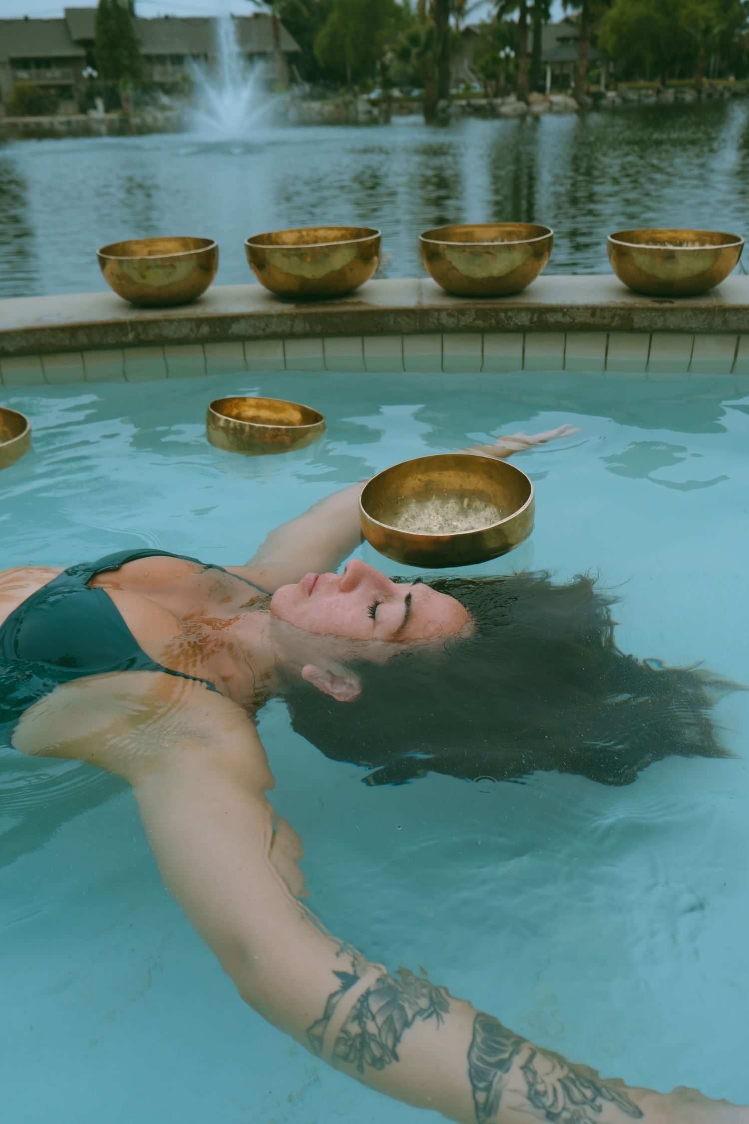 Person floating in pool with singing bowls and tattoo visible against tropical landscape.
