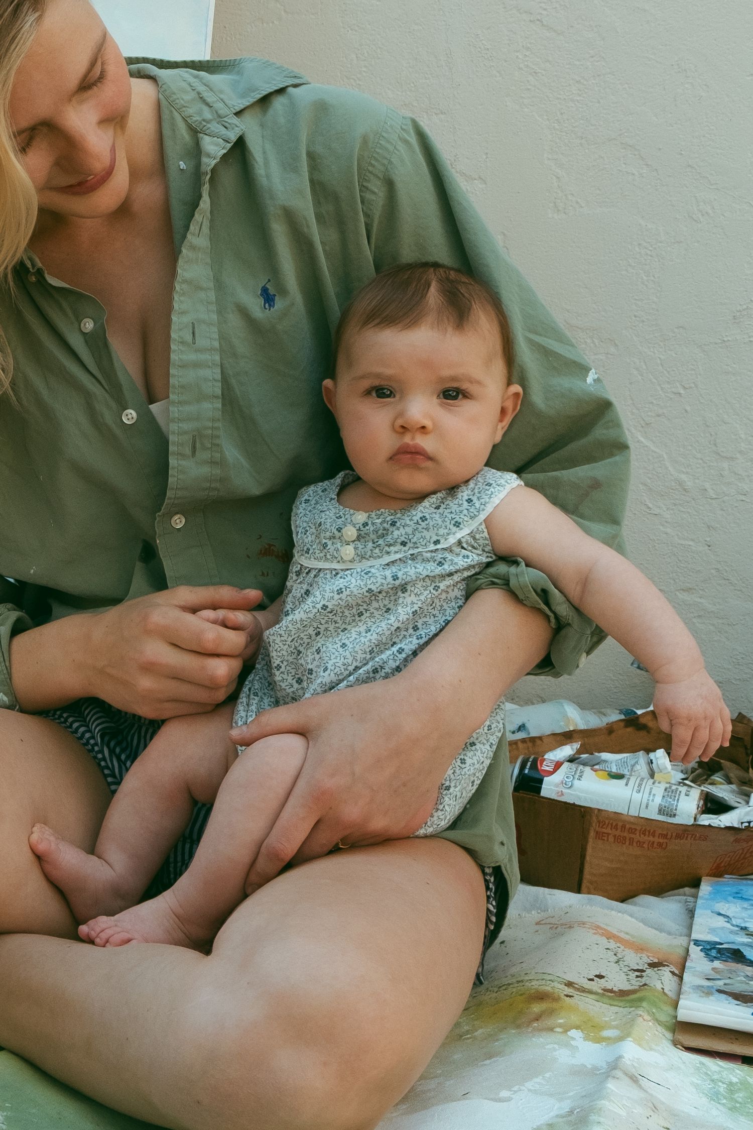 A baby wearing a floral dress sits comfortably on someone's lap in a casual outdoor setting.