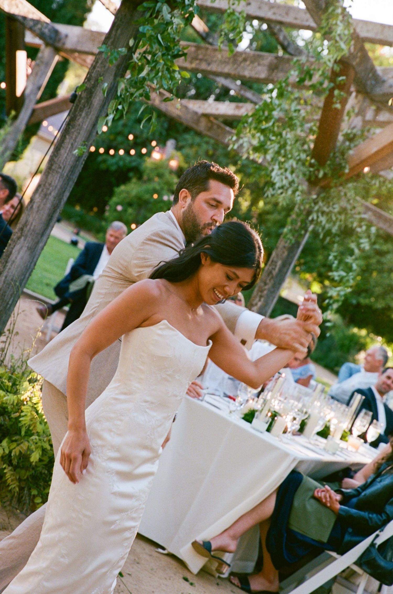 Bride and groom dancing joyfully under a rustic wooden pergola at an outdoor wedding reception.