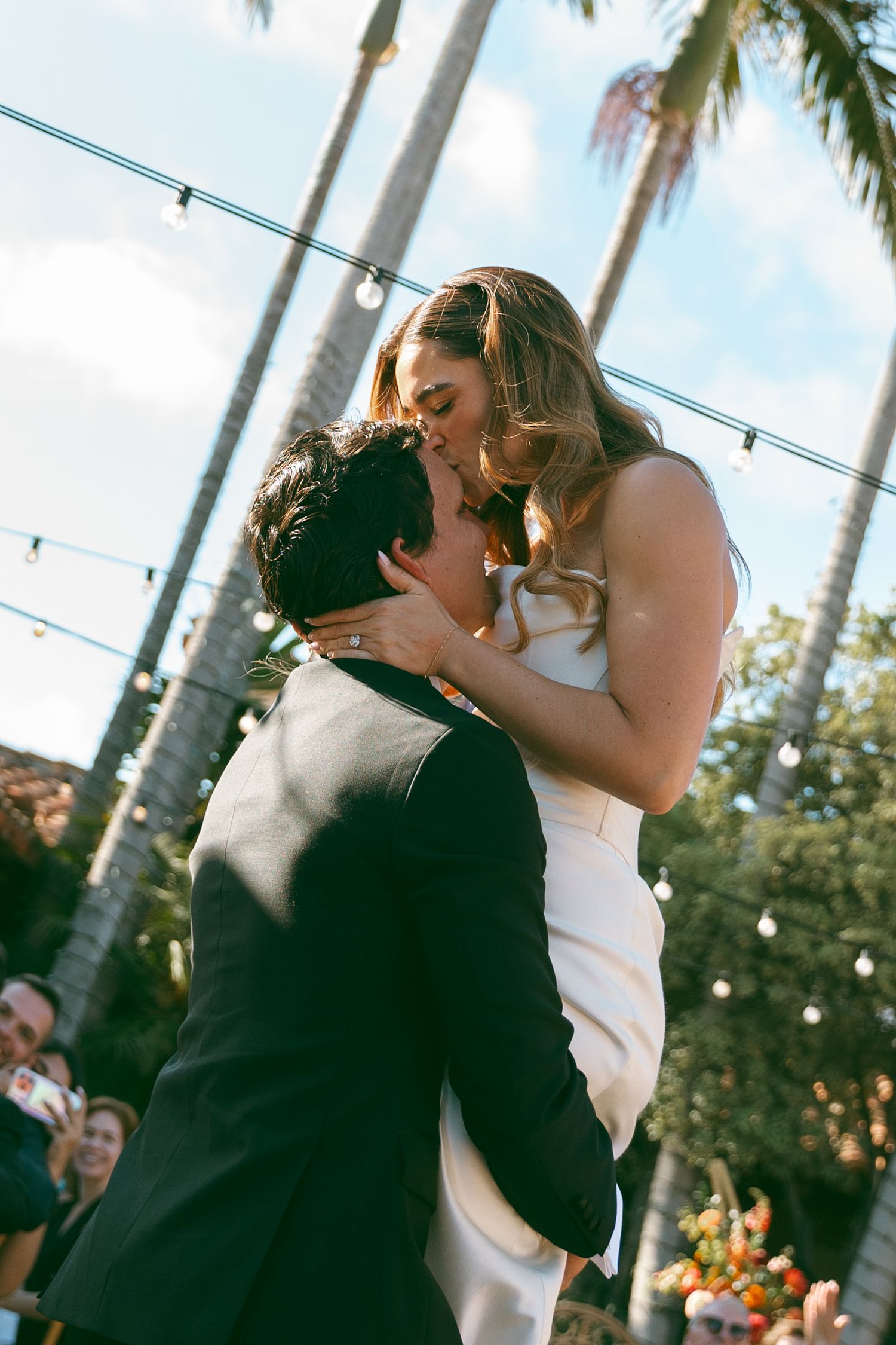 Groom lifting bride for a kiss outdoors at wedding ceremony with palm trees and string lights.