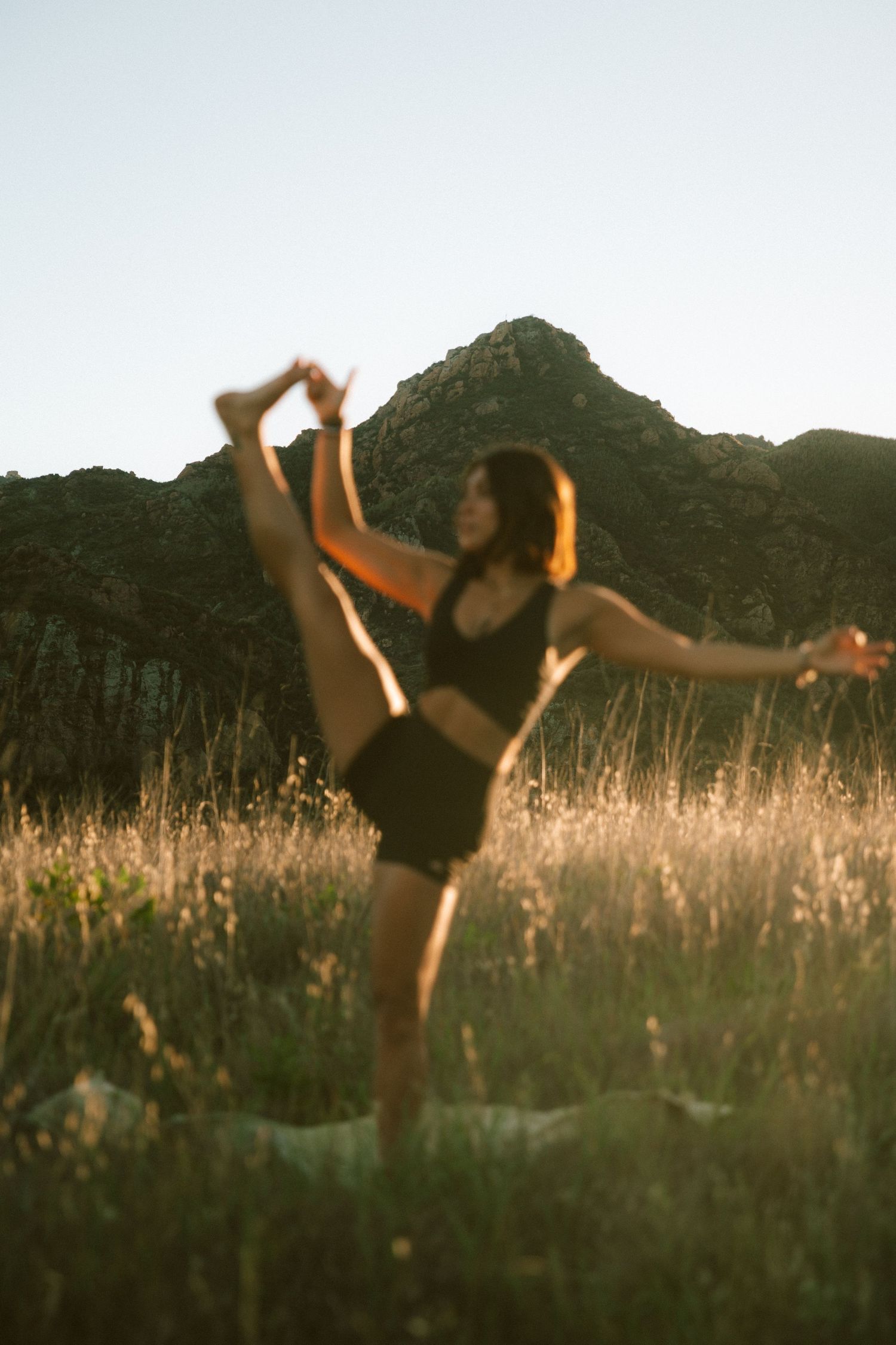 Silhouetted figure performing dance movements in a sunlit field with mountains in background.
