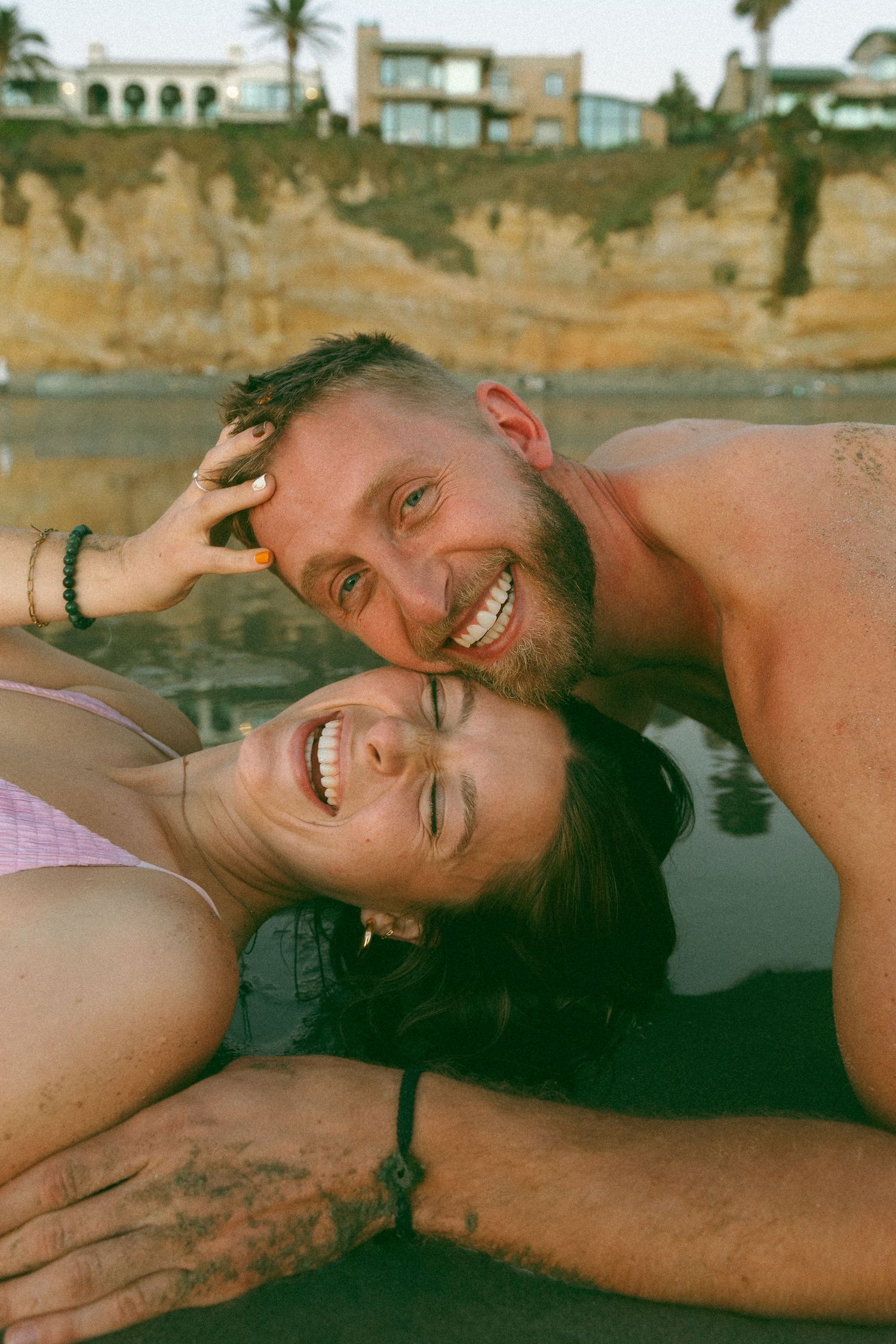 Couple laughing and embracing on a beach with coastal cliffs in the background.
