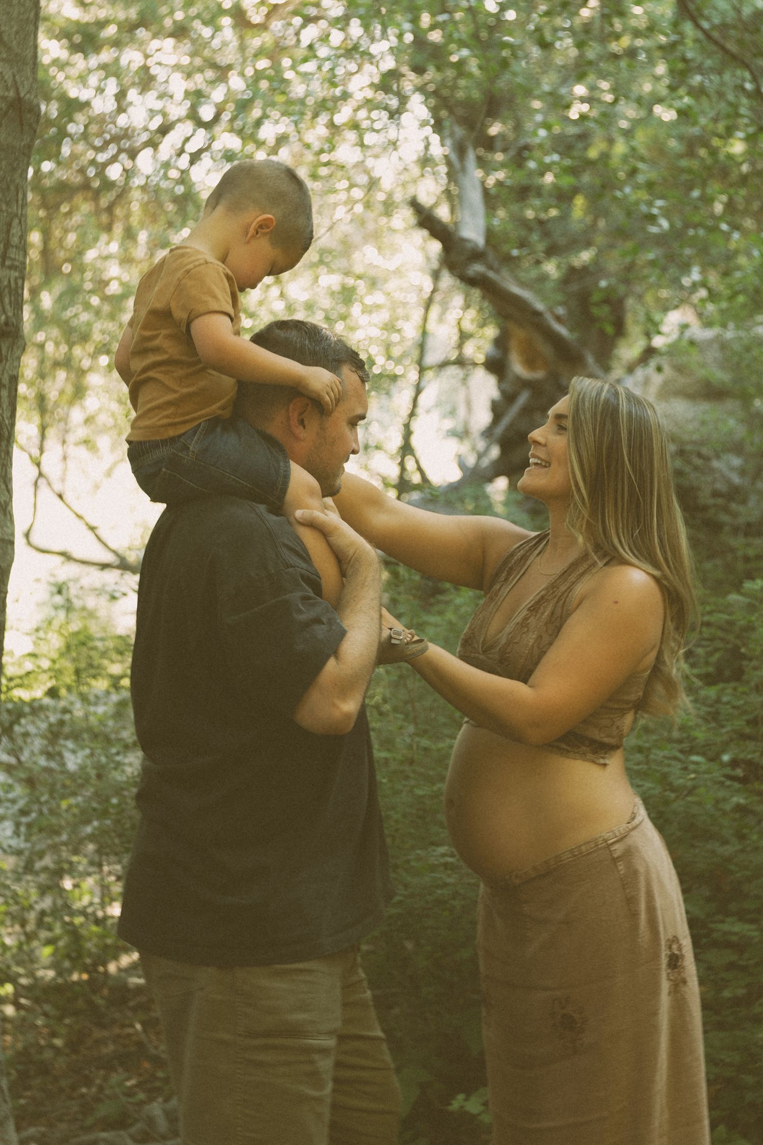 Candid family moment captured during outdoor maternity photoshoot in wooded setting.