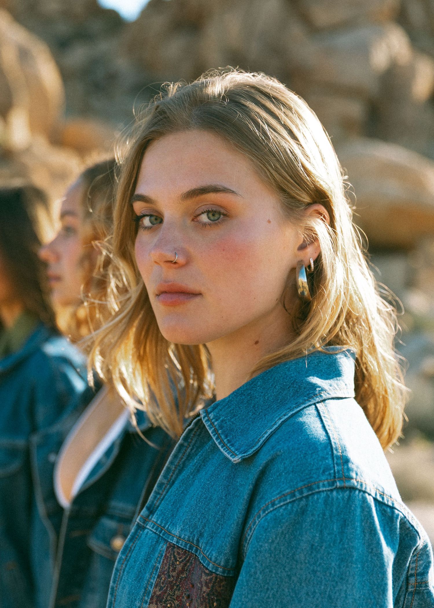 A person wearing a denim jacket poses outdoors in warm sunlight with a blurred background.