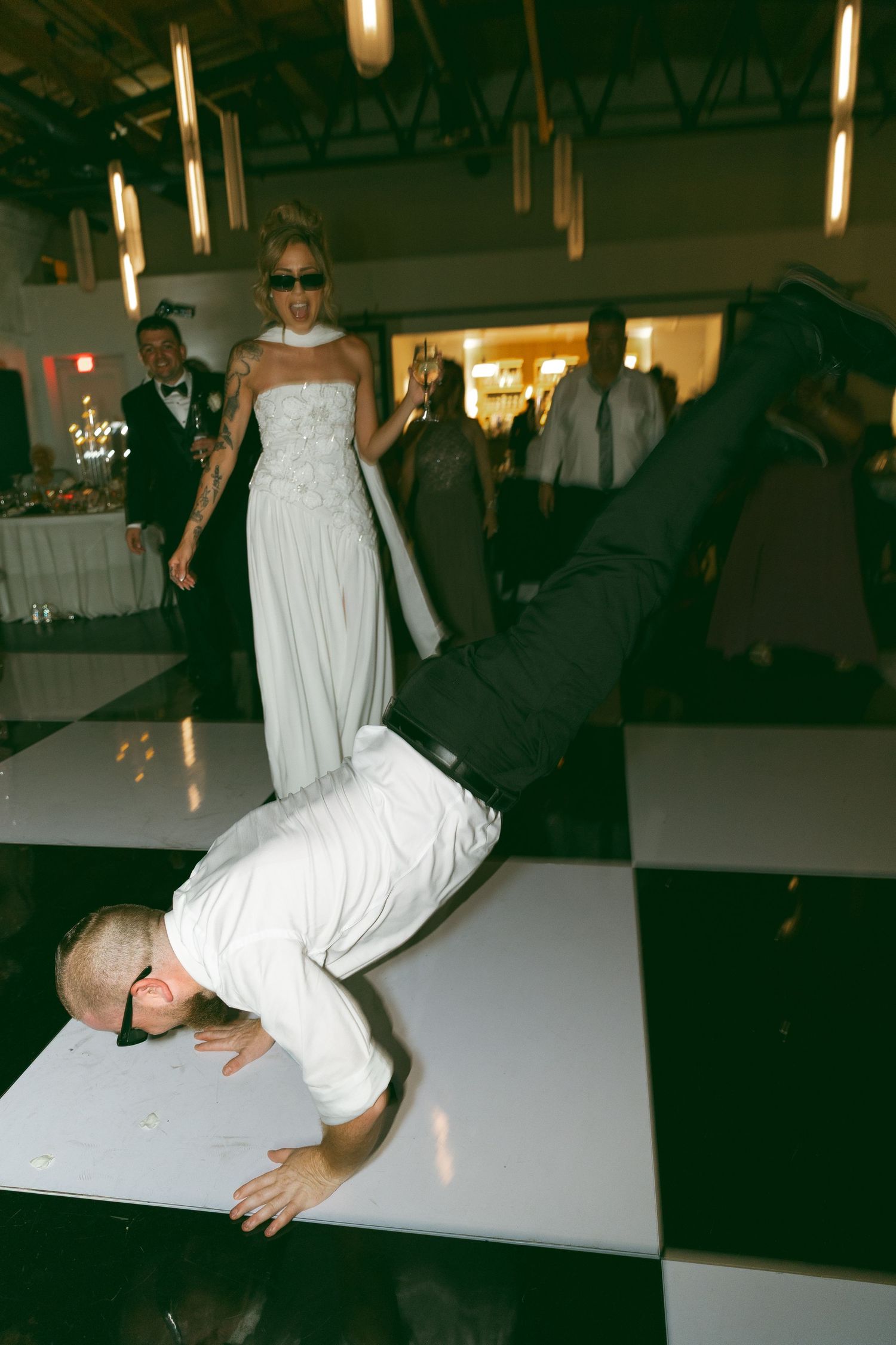Groom breakdancing on checkered dance floor at wedding reception while bride watches in white gown.