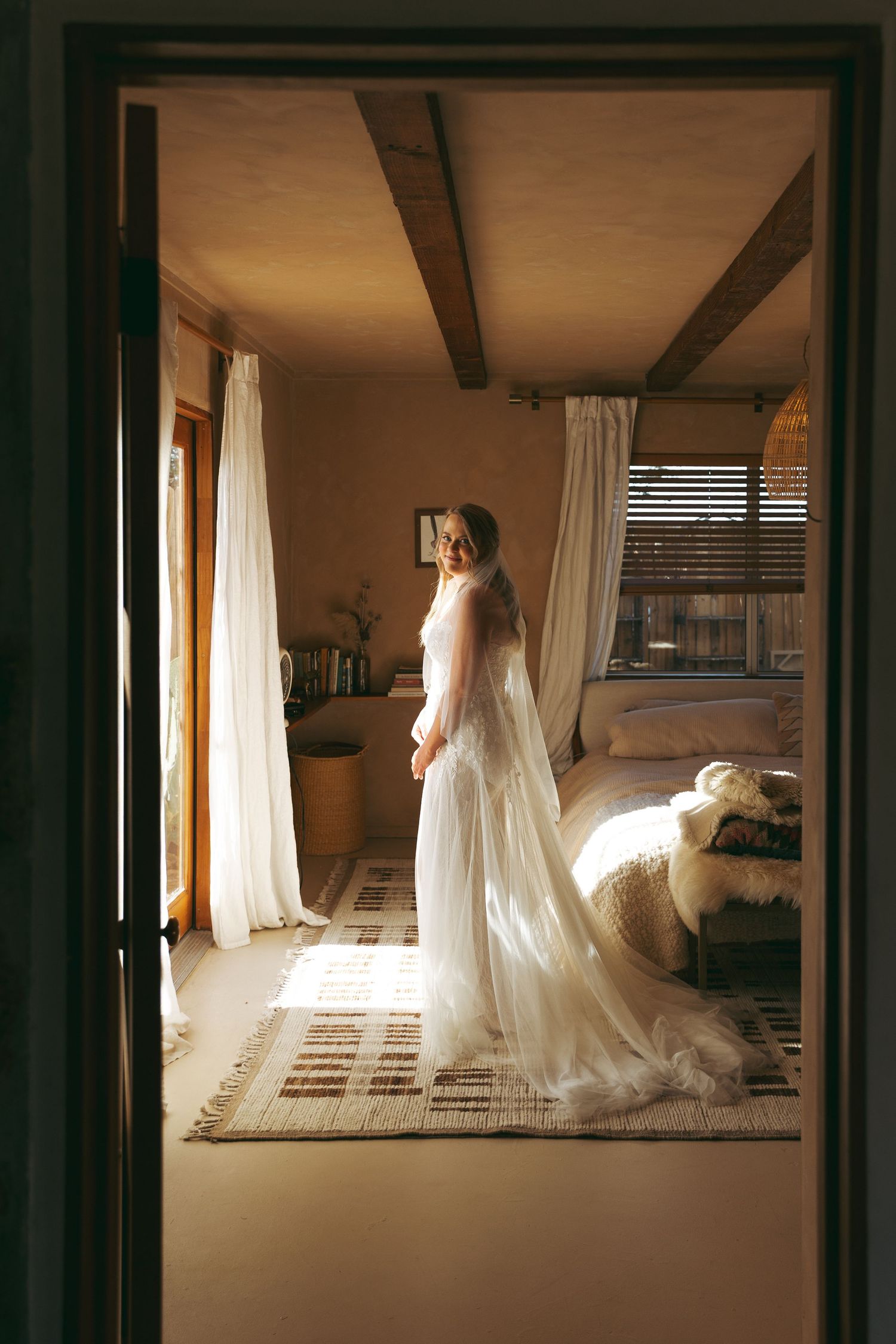 Bride in white wedding gown with long train standing by sunlit window in rustic bedroom.