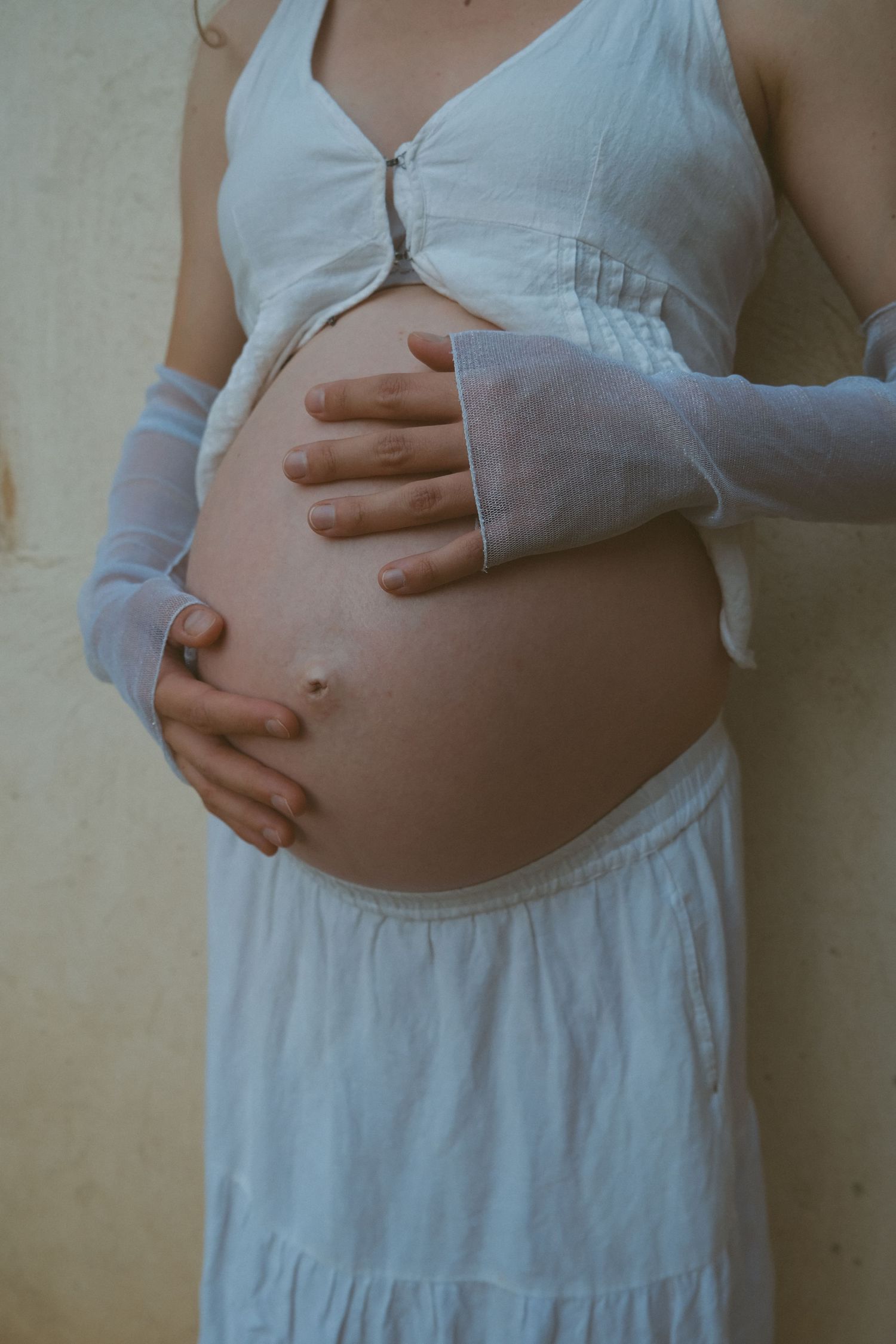 Hands gently cradle a pregnant belly while wearing a white dress and arm warmers.