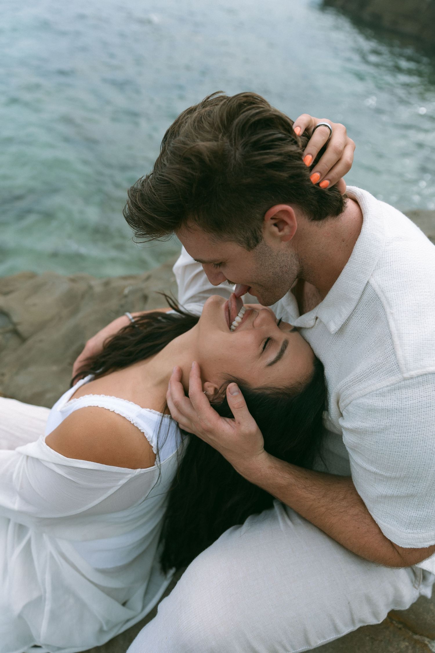 A couple dressed in white shares tender moments on a rocky coastline.