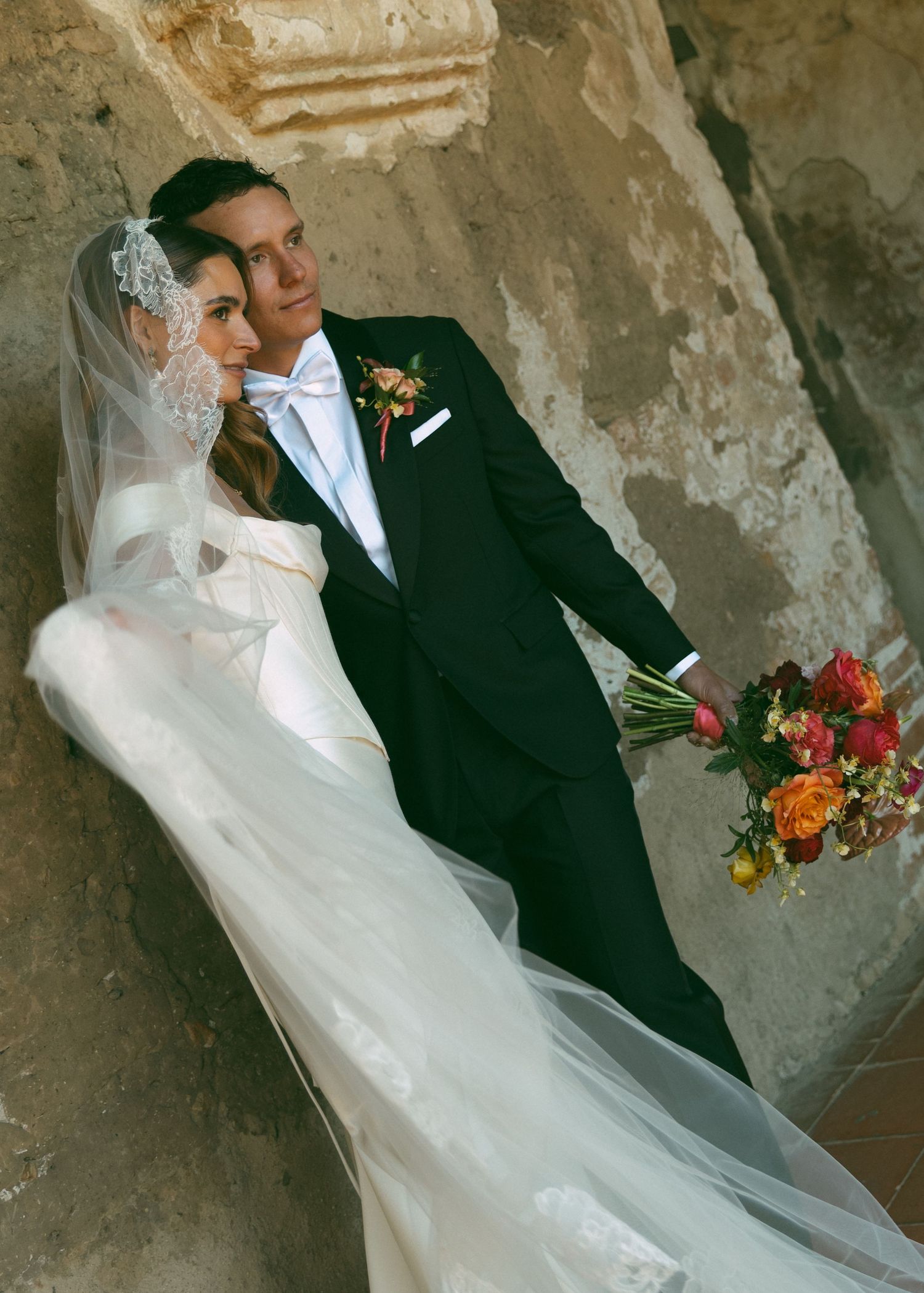 Bride in white gown and veil with groom in black tuxedo posing against stone wall holding colorful bouquet.