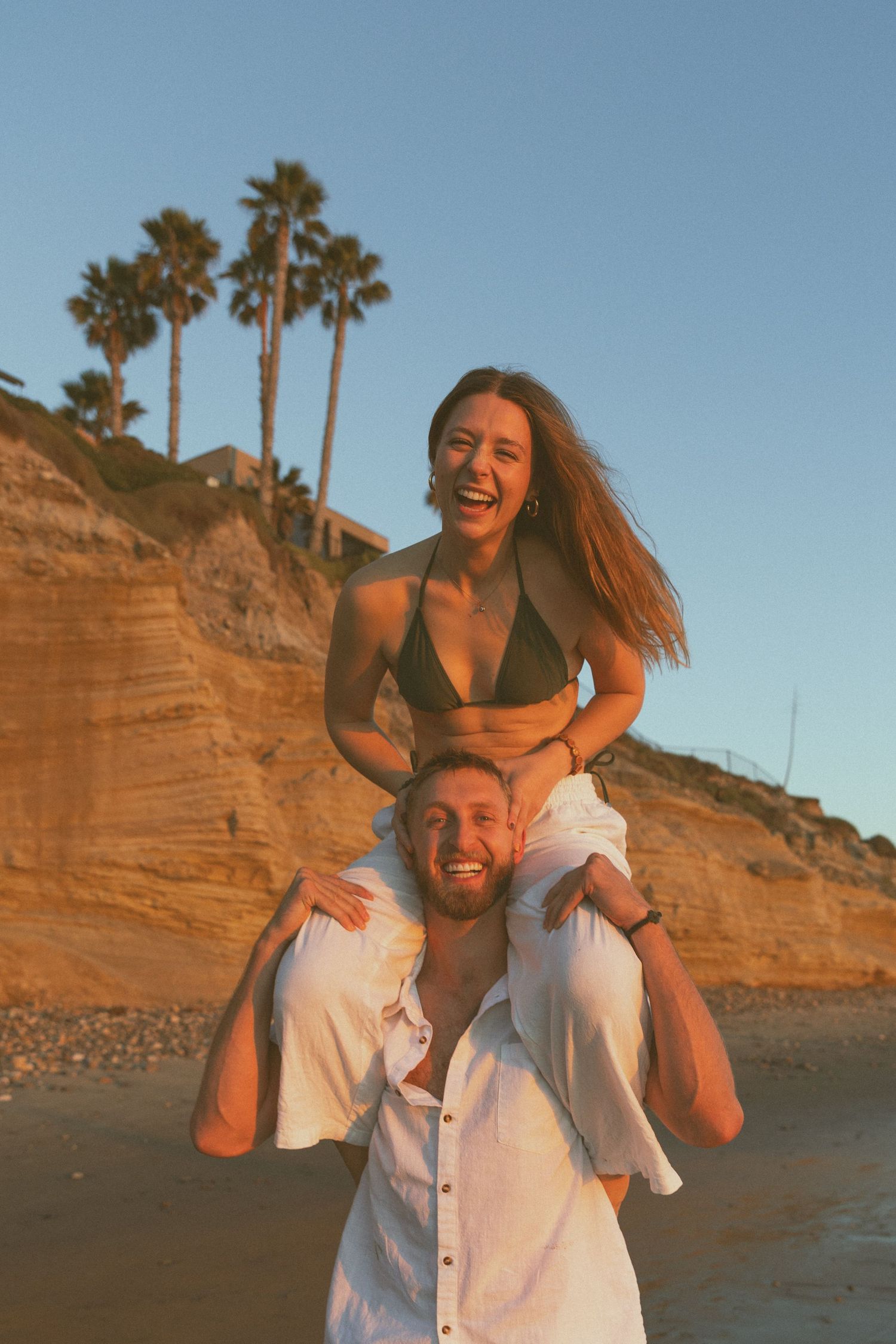 Playful piggyback ride photos on a beach with coastal cliffs in the background.
