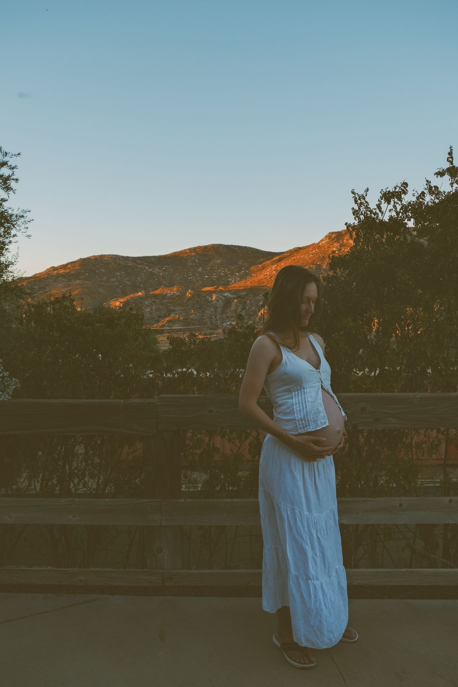 Maternity portrait in white dress against mountain backdrop at sunset.