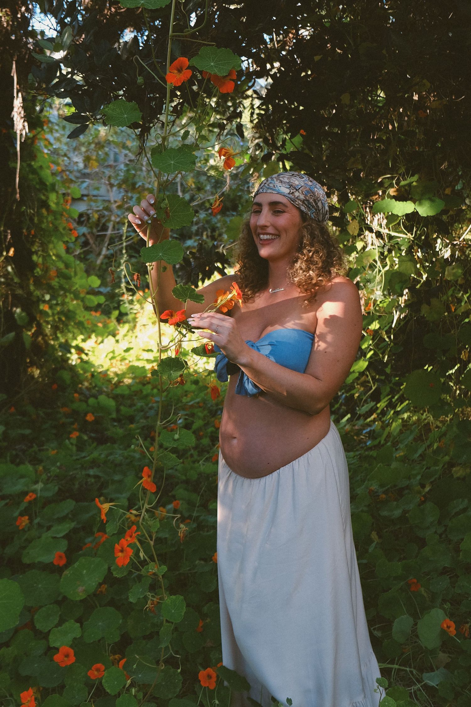 A candid garden maternity shoot with orange flowers and natural sunlight filtering through leaves.